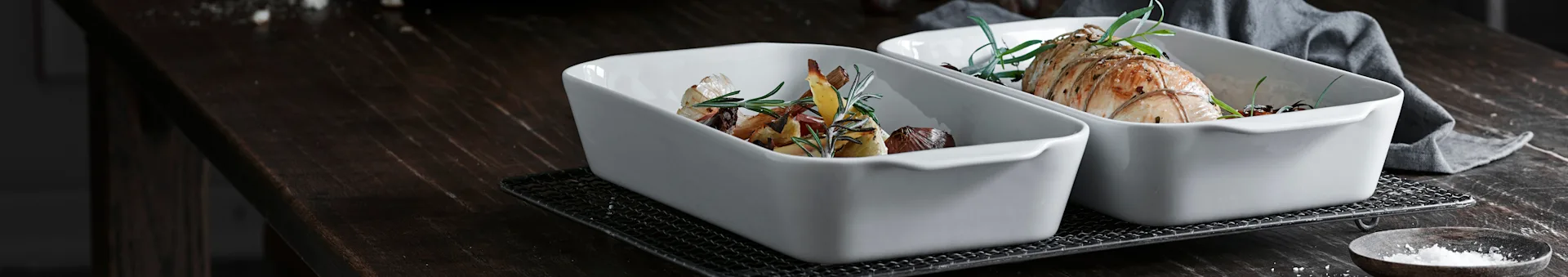 Two white baking dishes on a dark table: one with roasted vegetables and herbs, the other with a tied seasoned roast.