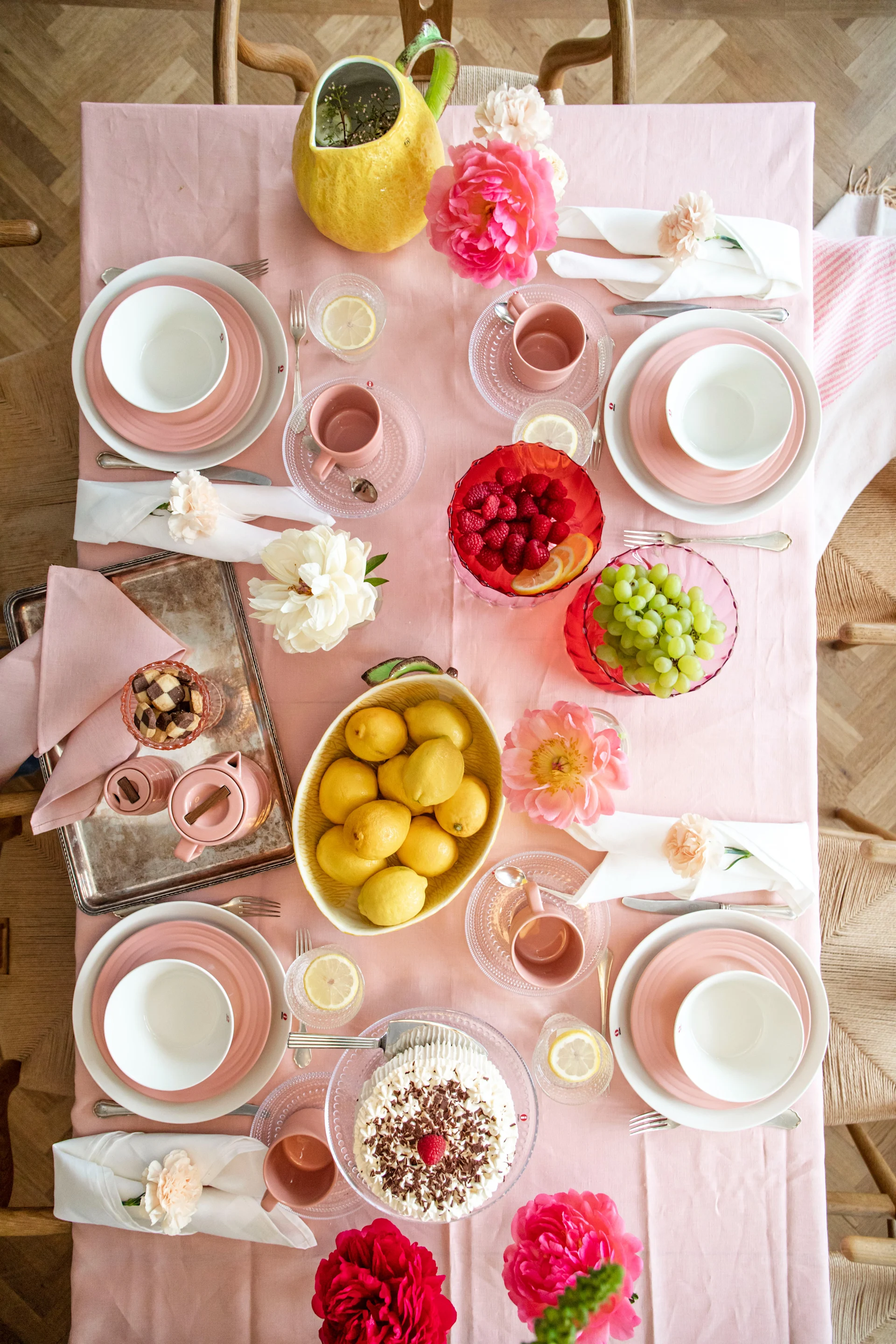 Overhead view of a dining table with pink tablecloth, white and pink place settings, fresh fruit, a cake, and pink flowers.