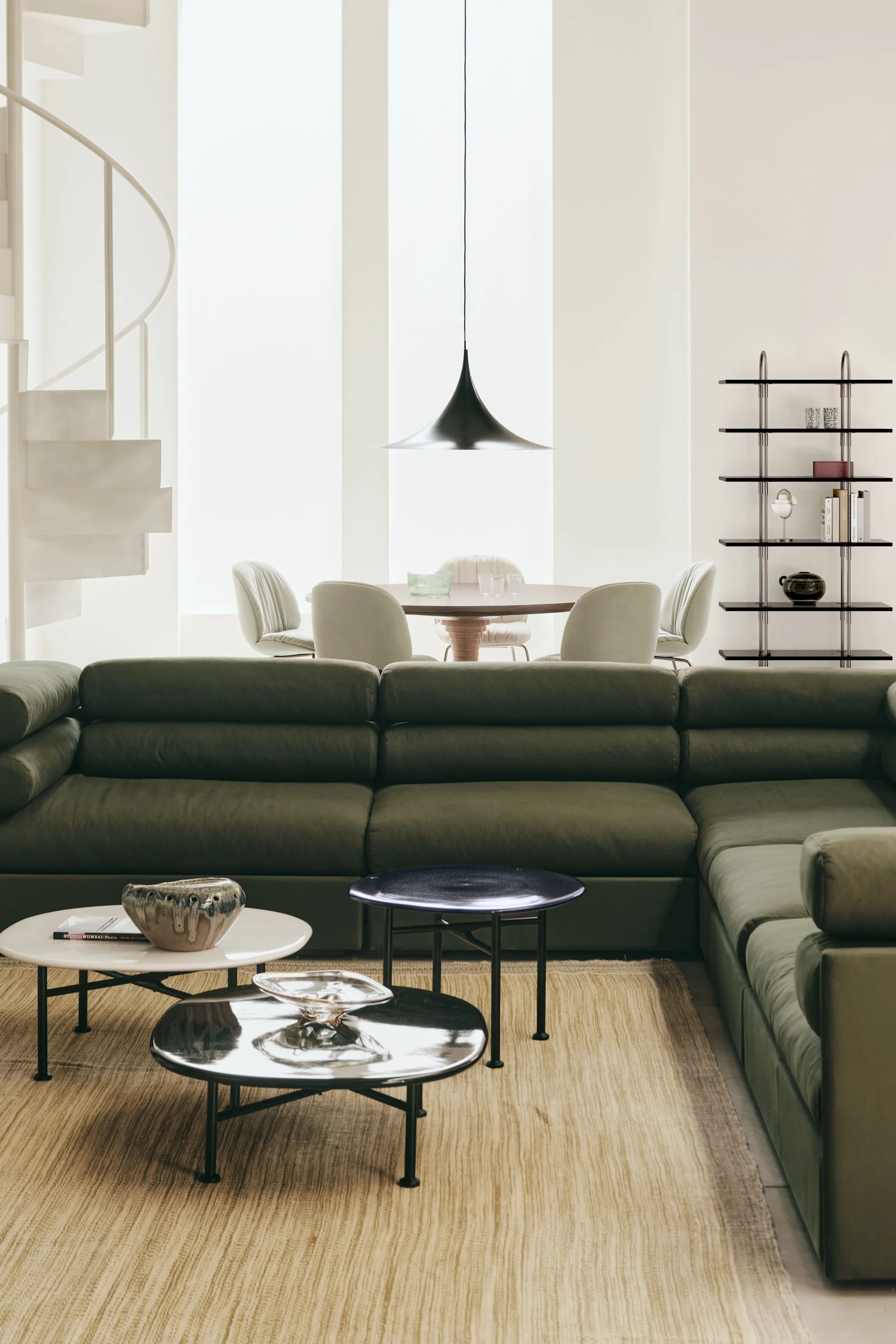 Modern living and dining space: olive green sofa, round tables, spiral staircase, dark shelving unit.