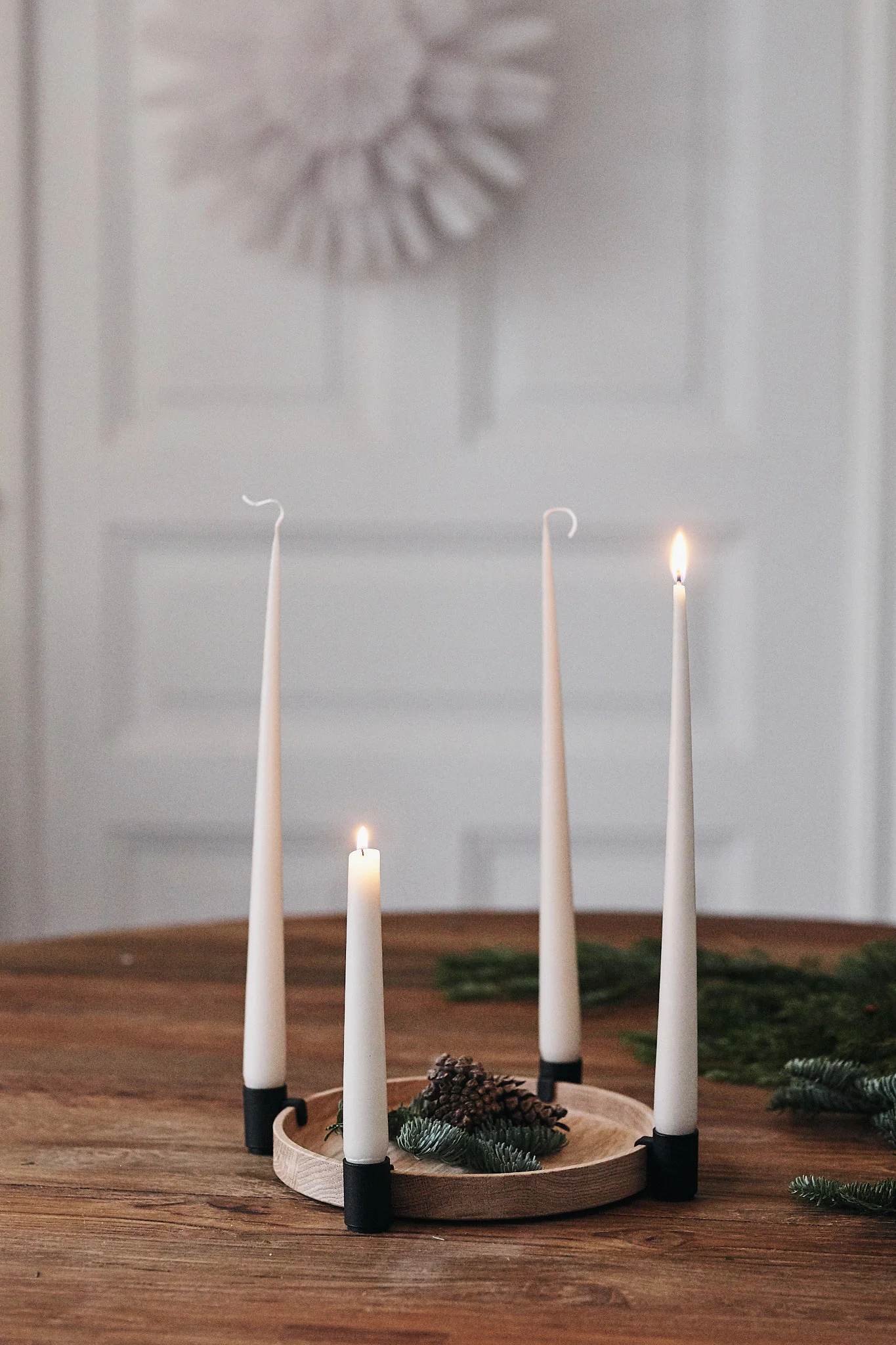 Four white taper candles, two lit, two unlit, on a wooden table with pine cones and branches.