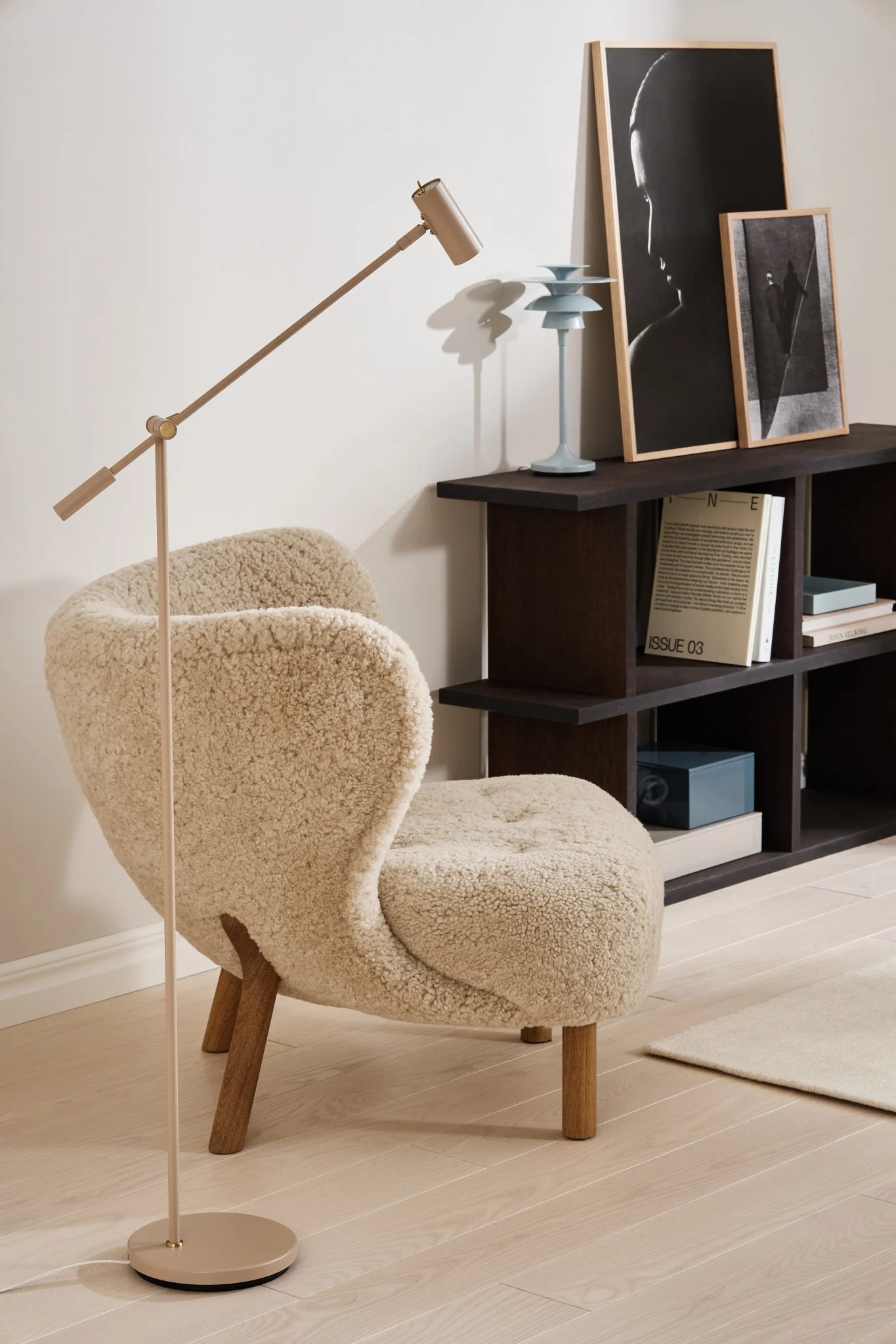 A cozy room with a cream fluffy armchair, a beige adjustable floor lamp, and a dark wooden shelf with art and books.