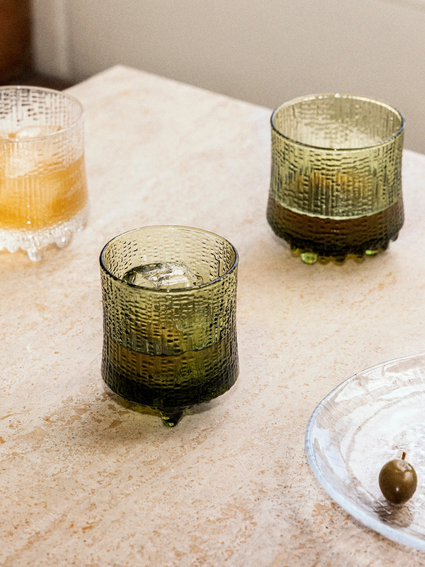 Three textured glasses with drinks and ice, two dark green and one clear, on a stone surface, next to a clear plate with an olive.