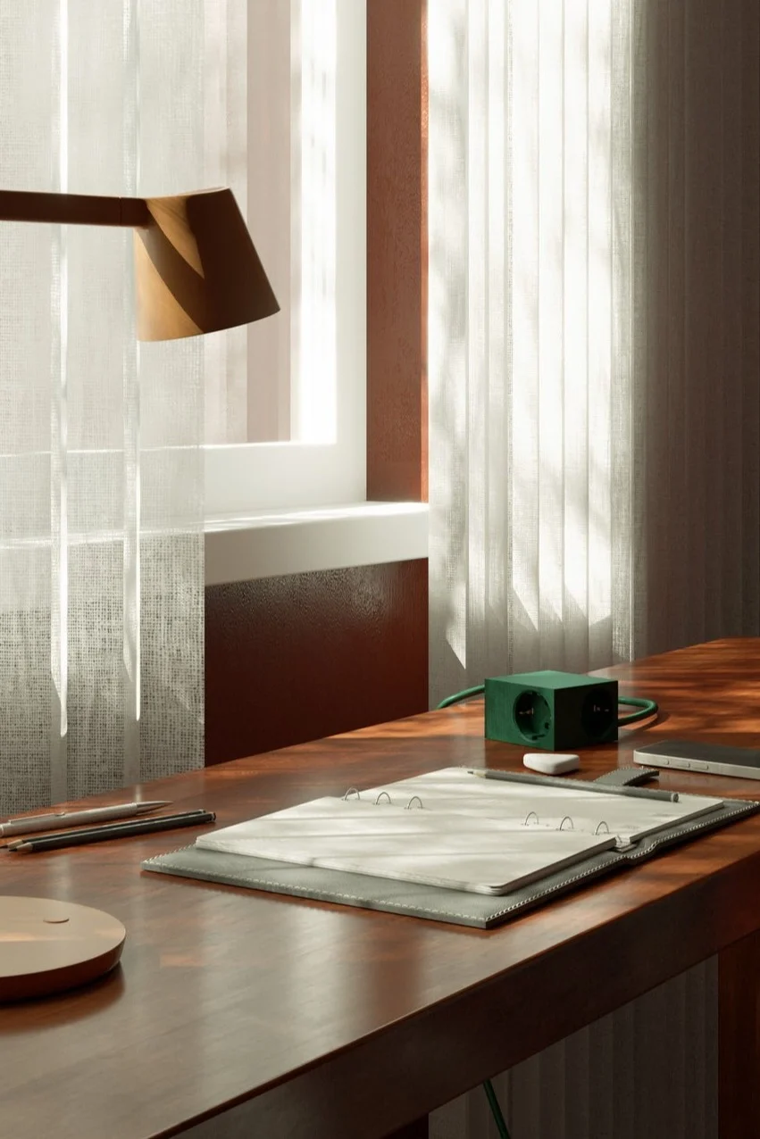 A tidy wooden desk with an open notebook, pencils, eraser, green cube-shaped power strip, and a lamp by a sunlit window.