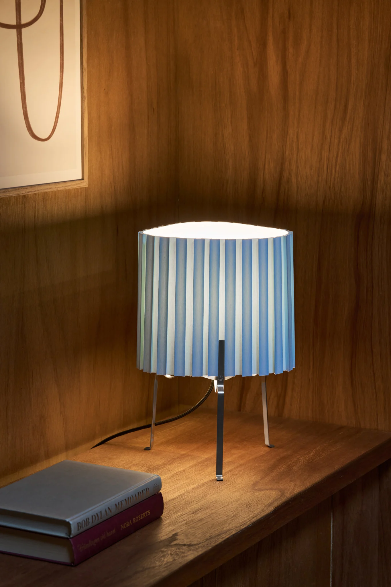 A table lamp with a blue and white striped pleated shade and metallic legs sits on a wooden shelf next to two books.