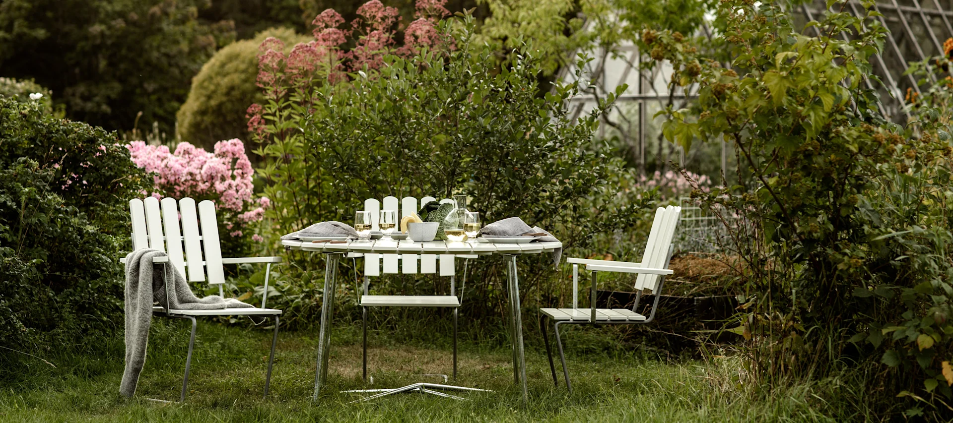 An outdoor dining setting with a round white slatted table and chairs set in a lush garden with pink flowers and a greenhouse.