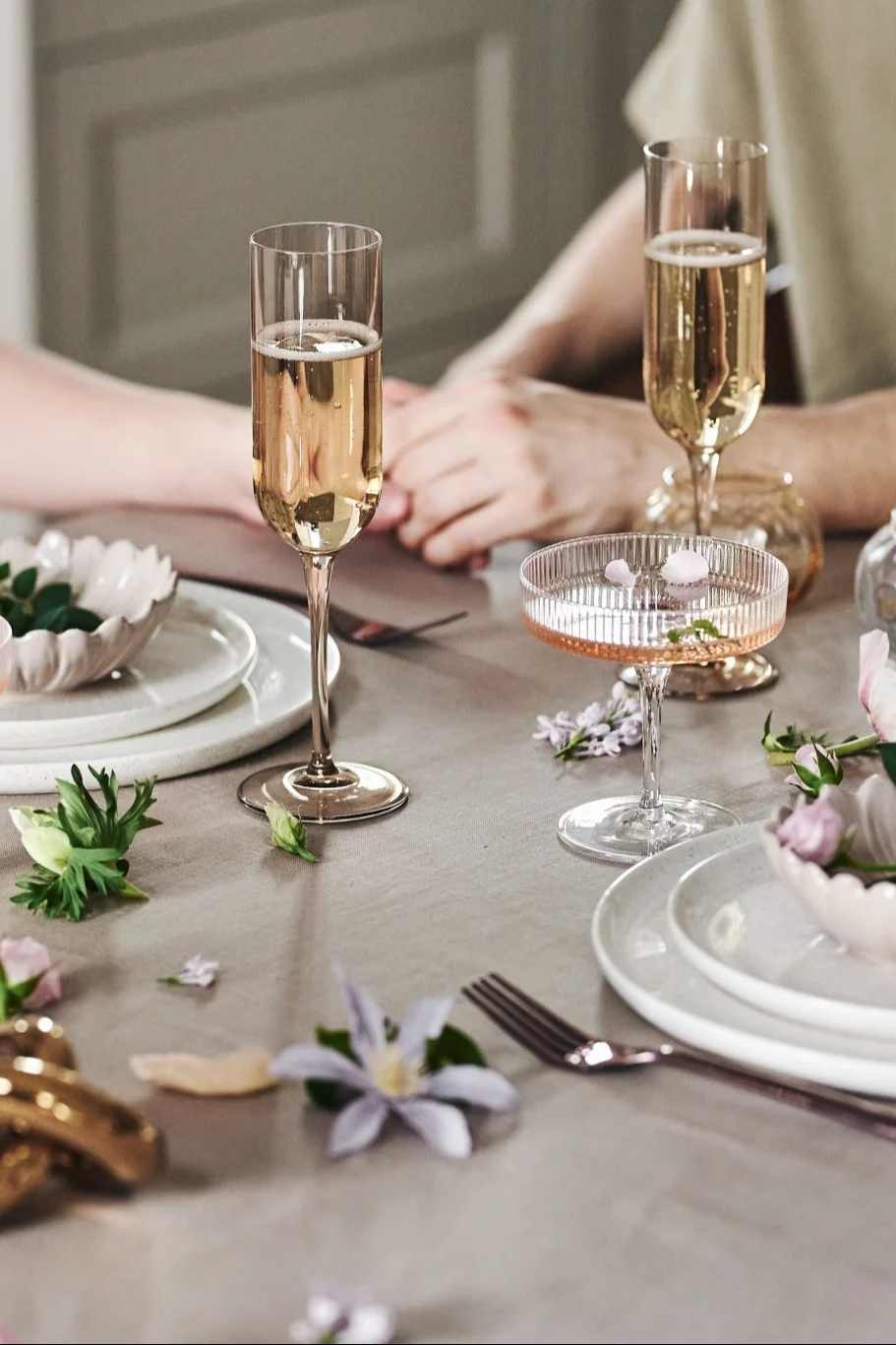 You can also decorate the table in a playful way for your romantic meal at home. Here you can see white crockery, simple glasses and flowers on a beige-coloured tablecloth.
