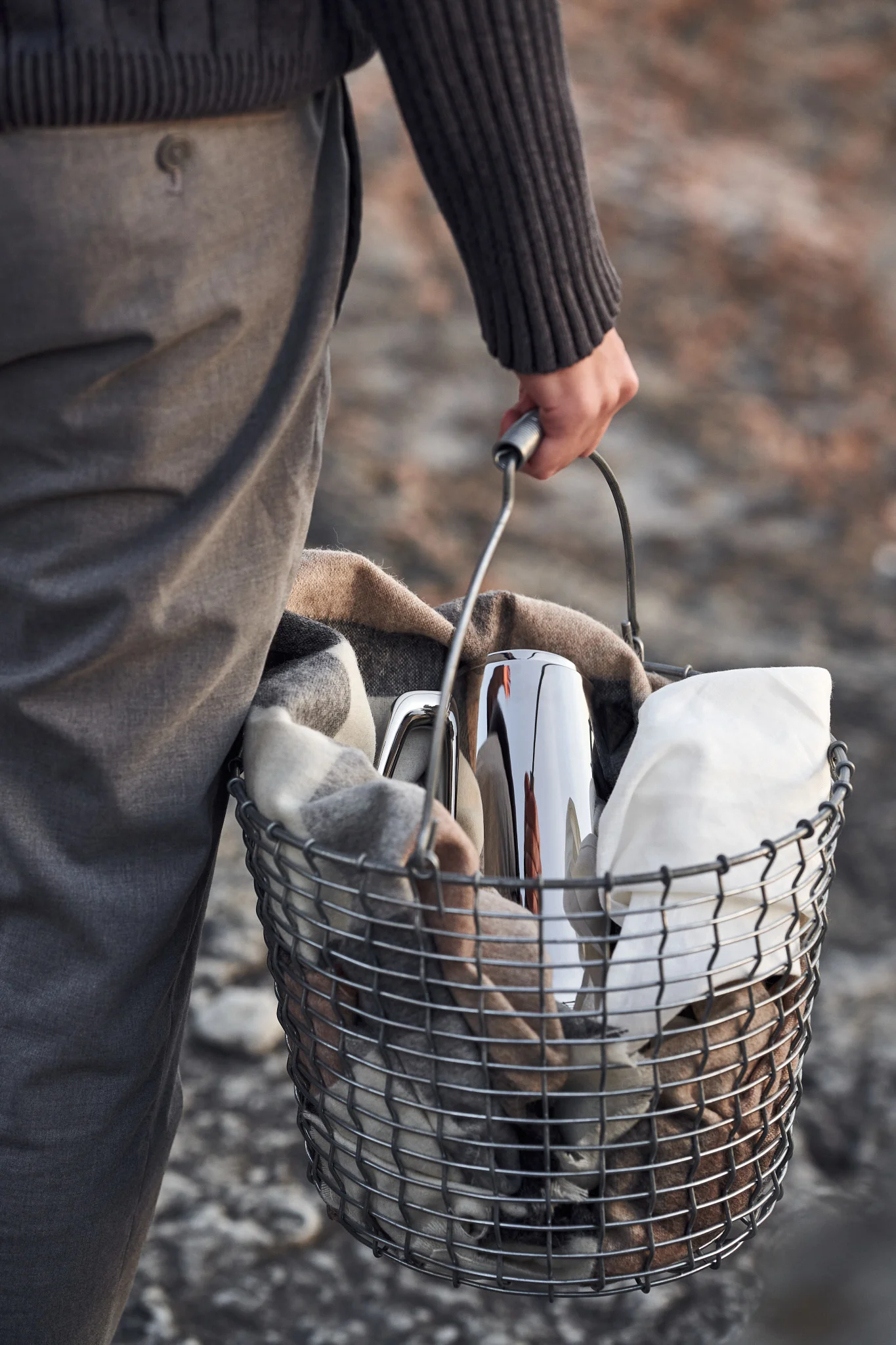 Person carrying a metal basket with a plaid blanket, a silver thermal carafe, and a white cloth.