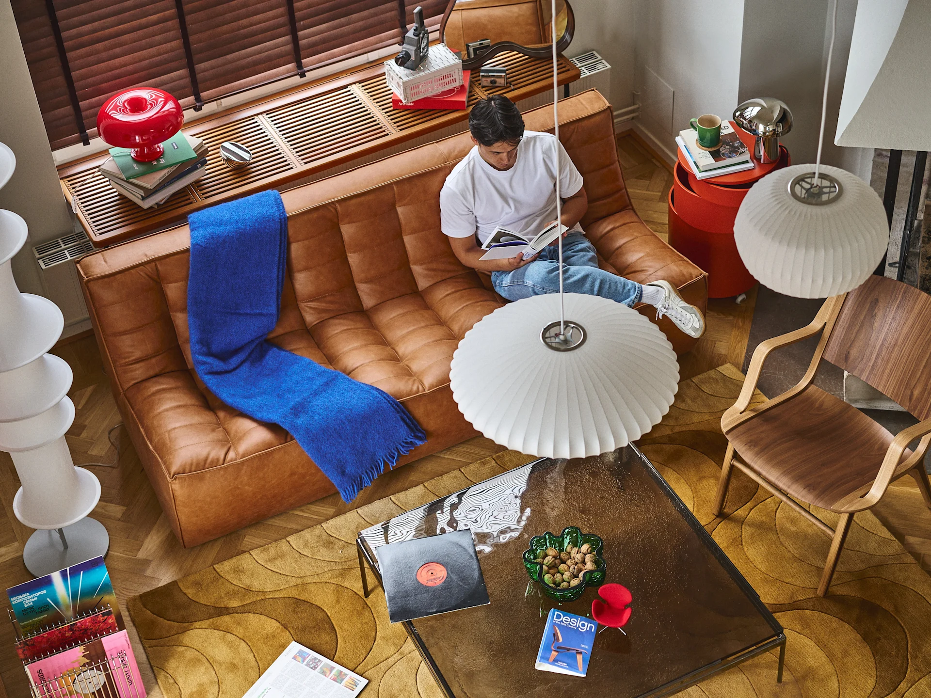 An overhead view of a man reading on a brown leather sofa, surrounded by design objects including a red lamp, records, and a patterned rug.