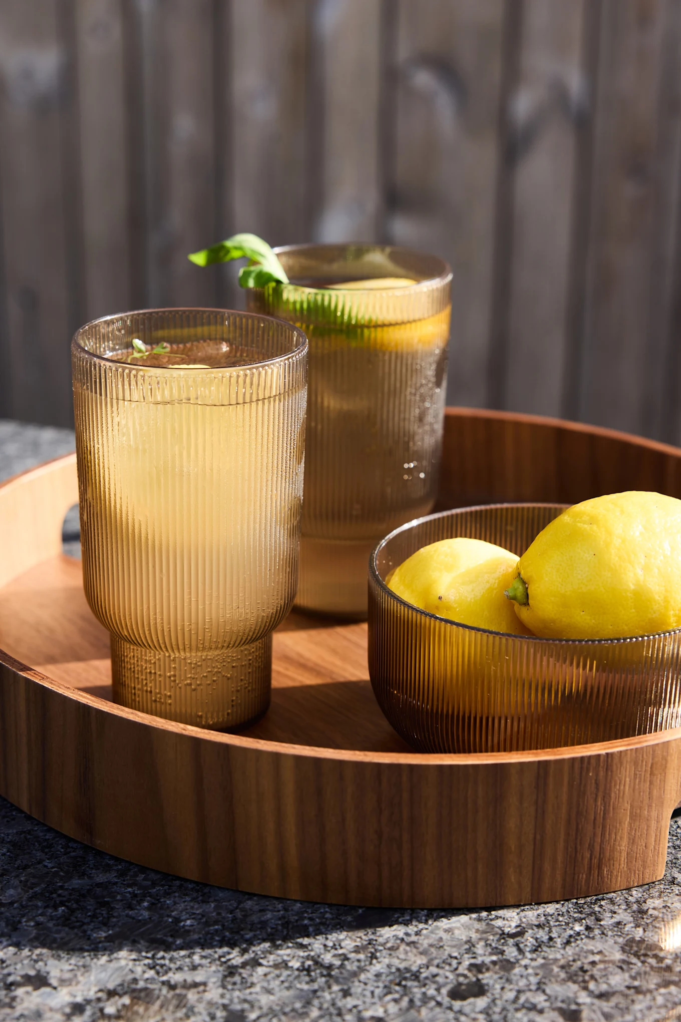 Two ribbed amber glasses with lemonade and a bowl of lemons on a wooden tray outdoors.