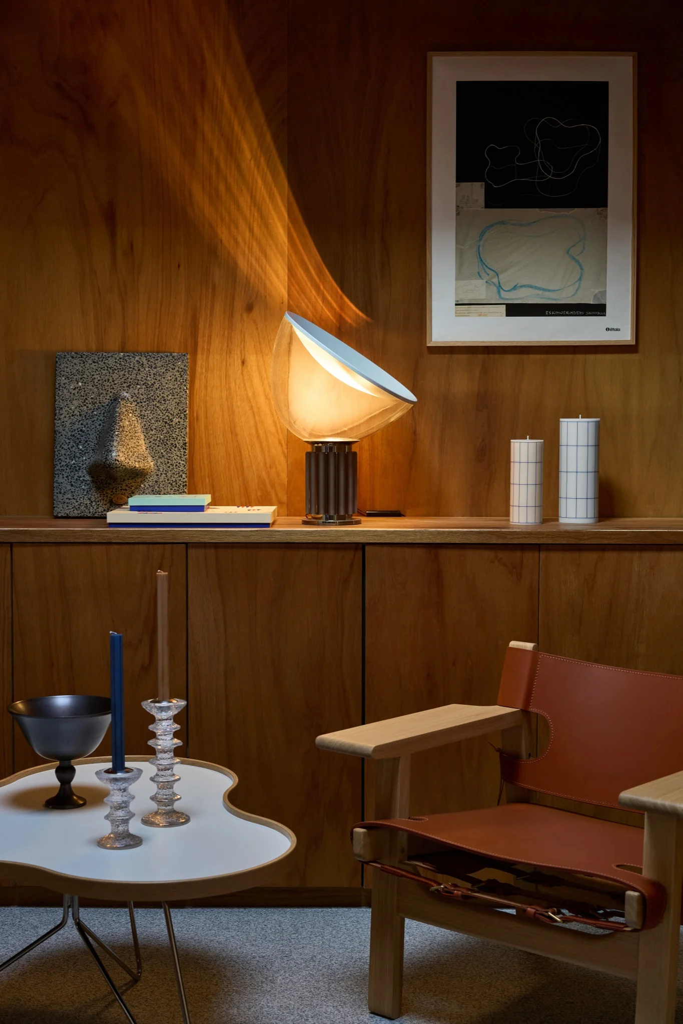 Wood-paneled room with a glowing lamp, framed art, grid candles on a shelf, a brown leather chair, and a white side table.