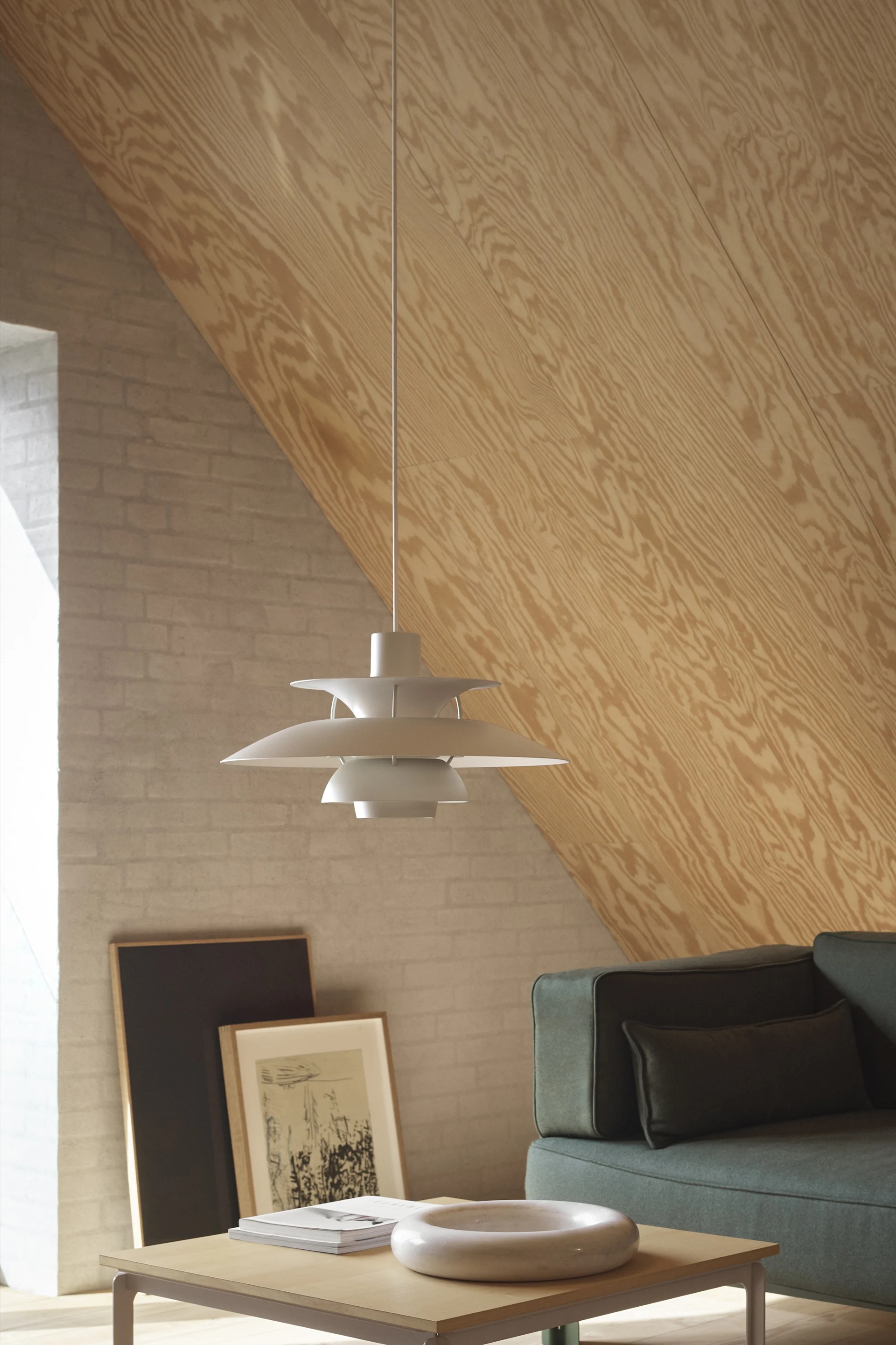 A light grey tiered pendant lamp hangs in a room with a sloped wooden ceiling, a brick wall, and a dark green sofa.