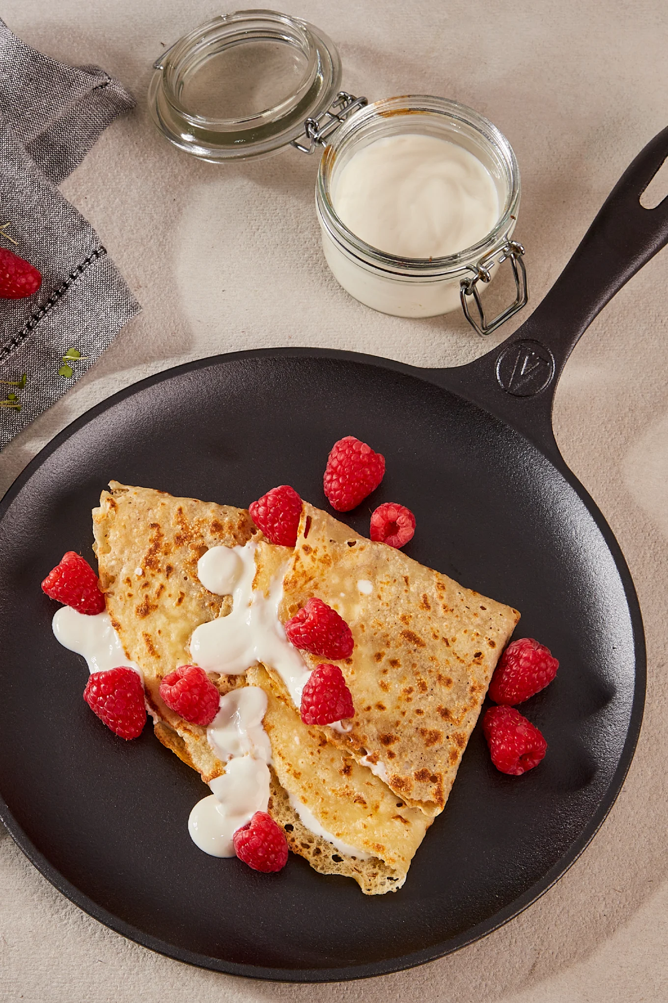 Two crepes with cream and fresh raspberries in a black pan, with a jar of cream and napkin in the background.
