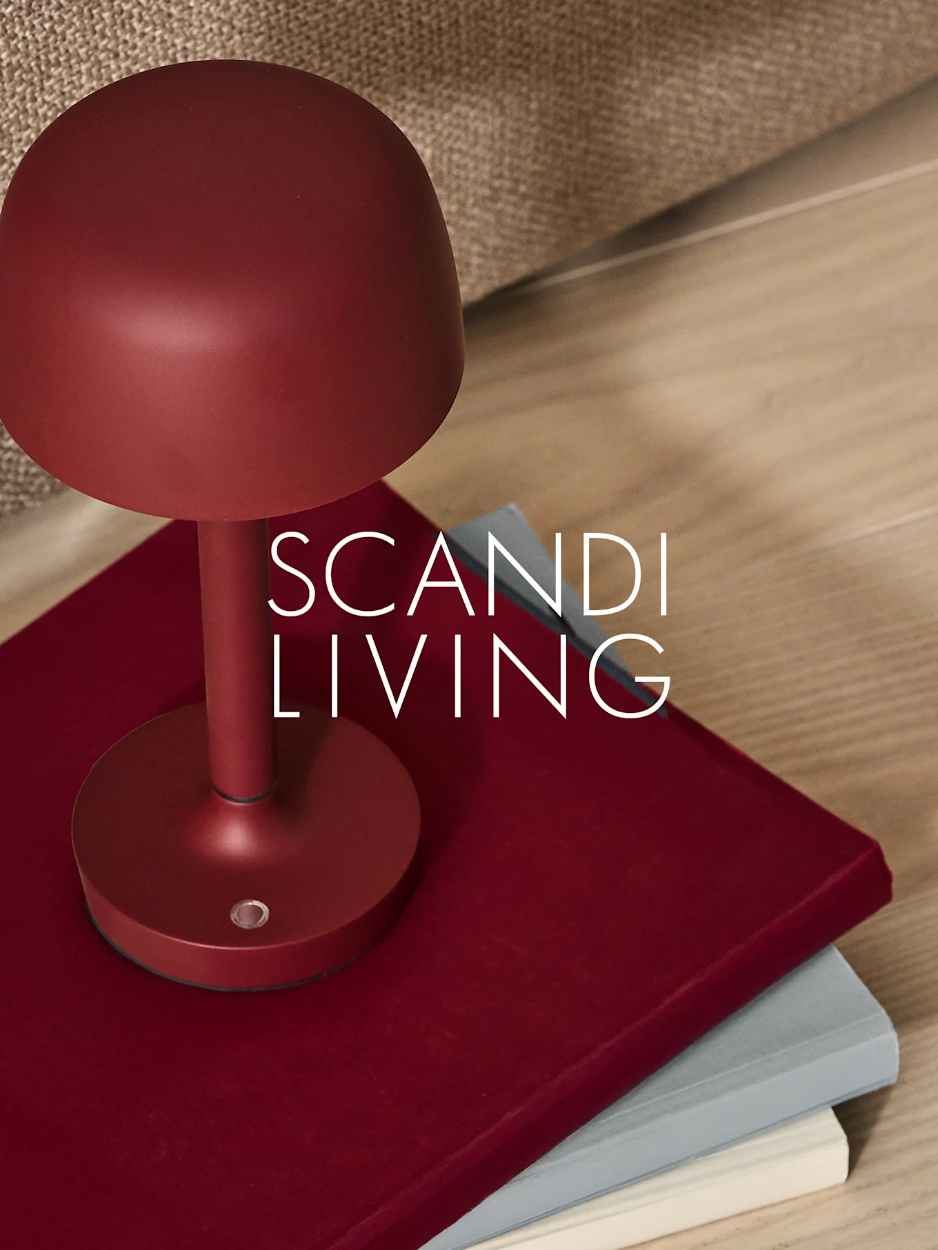 A dark red mushroom-shaped table lamp sits on two books, with a light brown fabric and wooden floor in the background.