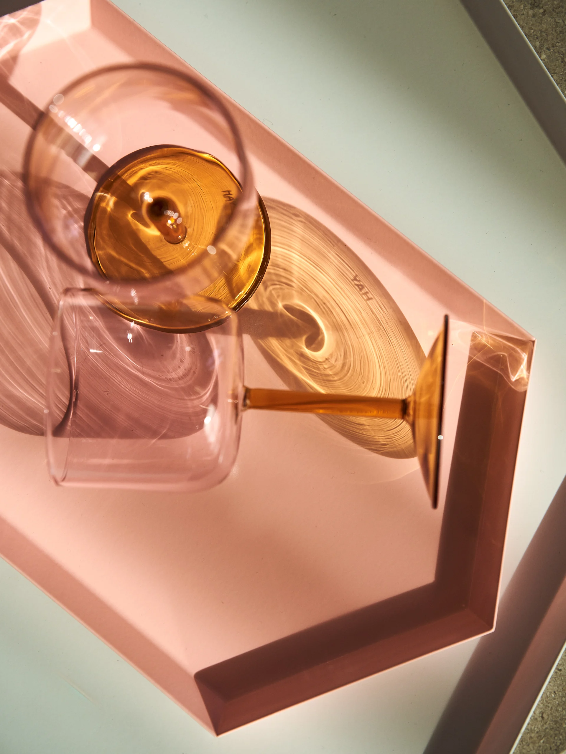 Two wine glasses, one amber and one clear, casting dynamic shadows on a pink tray and light surface.