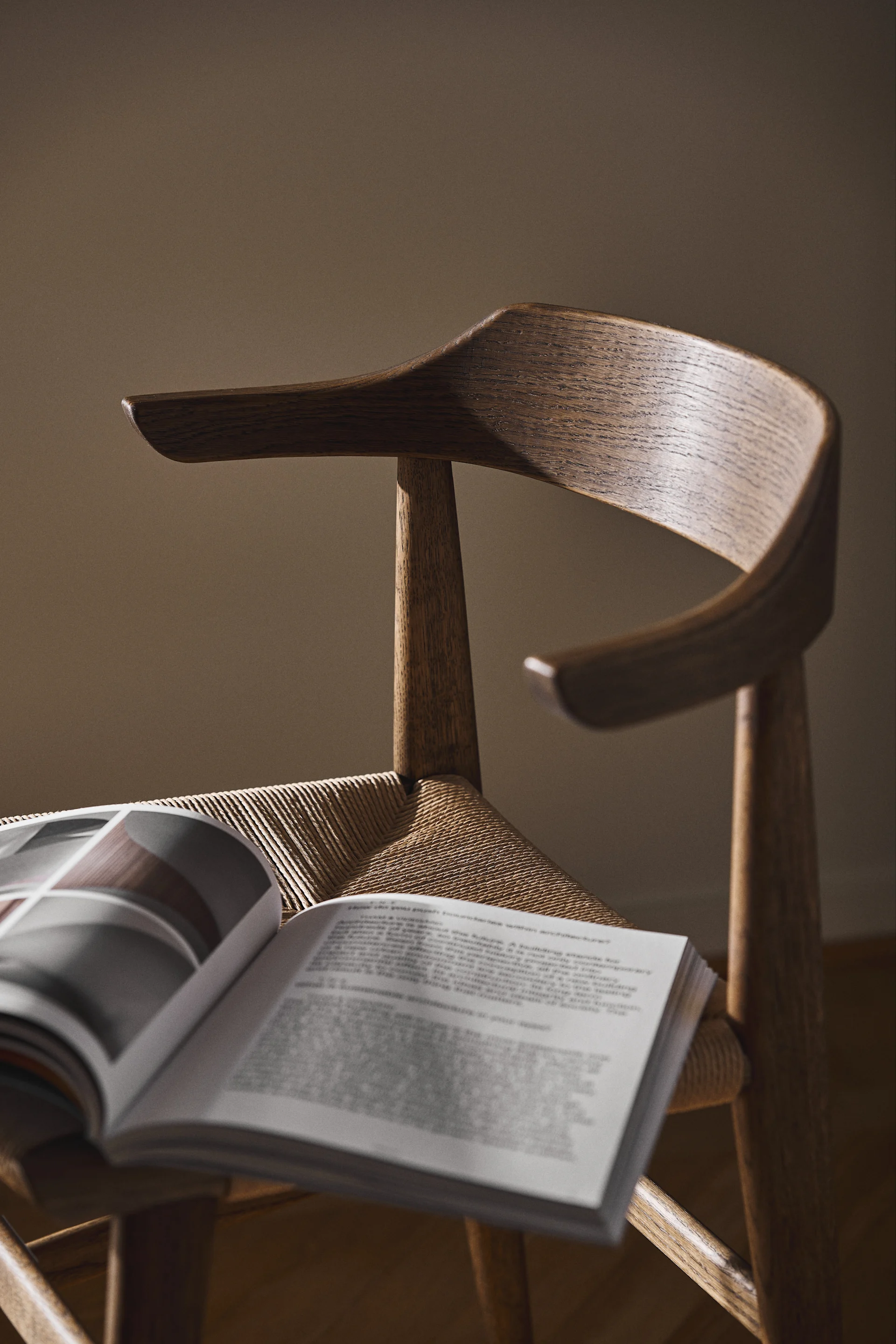 An open book rests on the woven seat of a wooden chair with a curved backrest, in soft light.