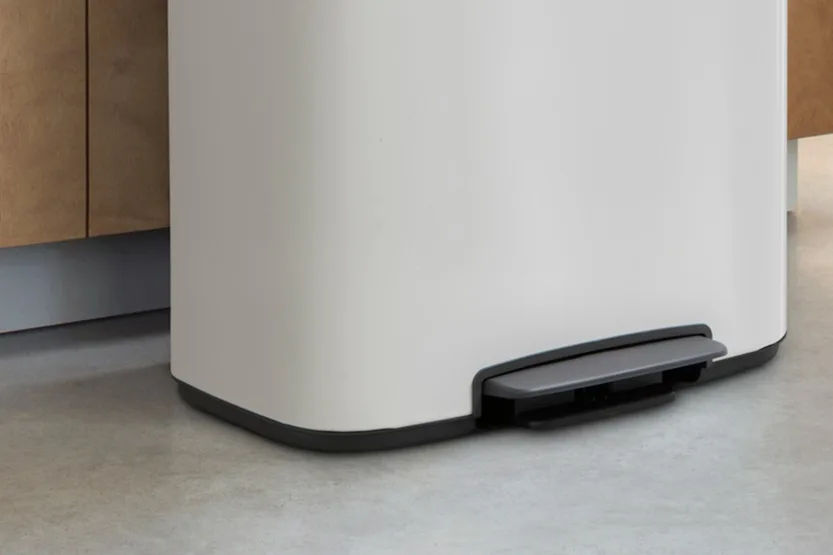 Bottom section of a light grey rectangular pedal bin with a dark grey pedal on a concrete floor next to a wooden cabinet.