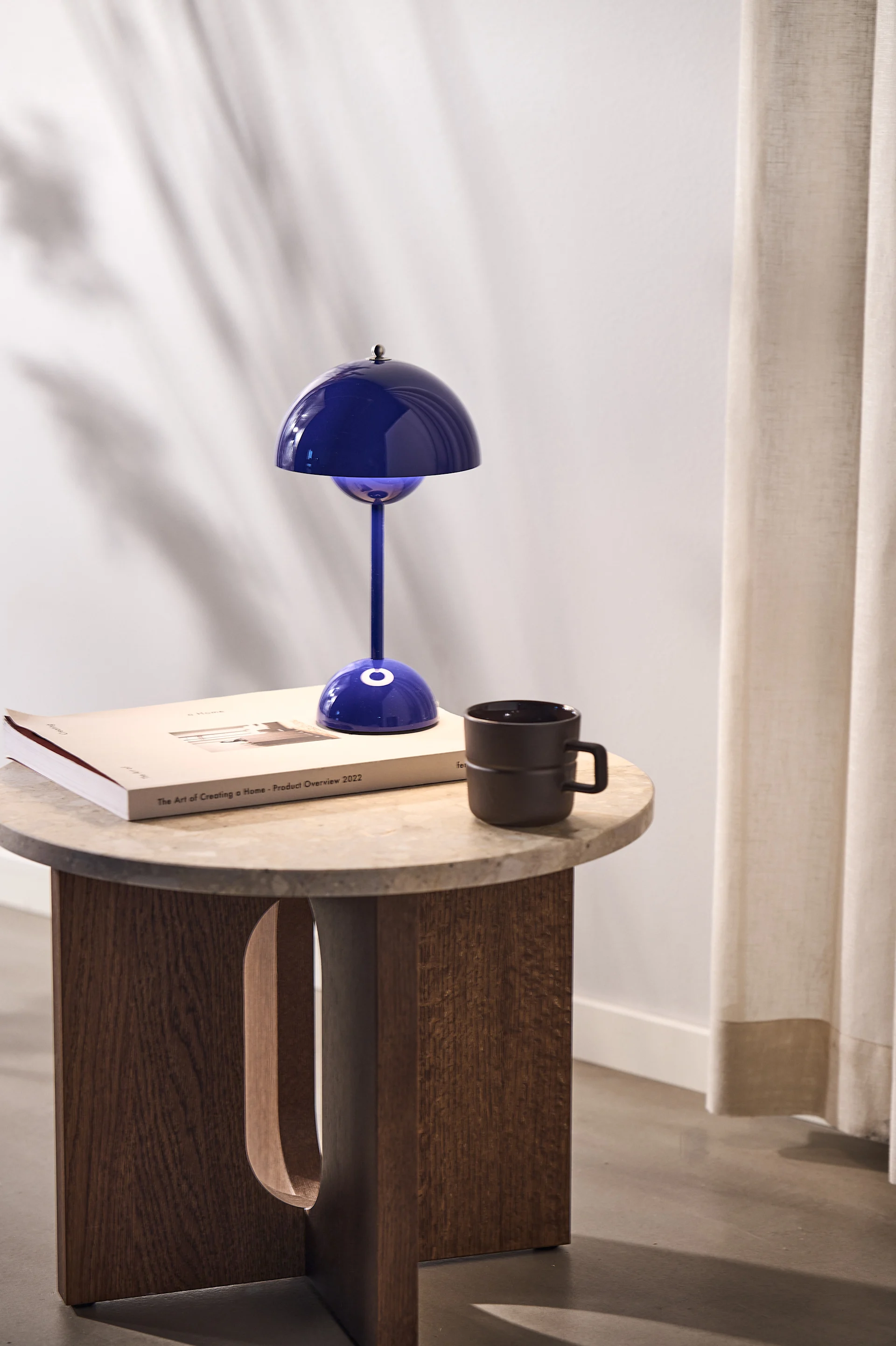 A blue mushroom table lamp, a book, and a dark mug on a round table with a wooden base, beside a curtain.