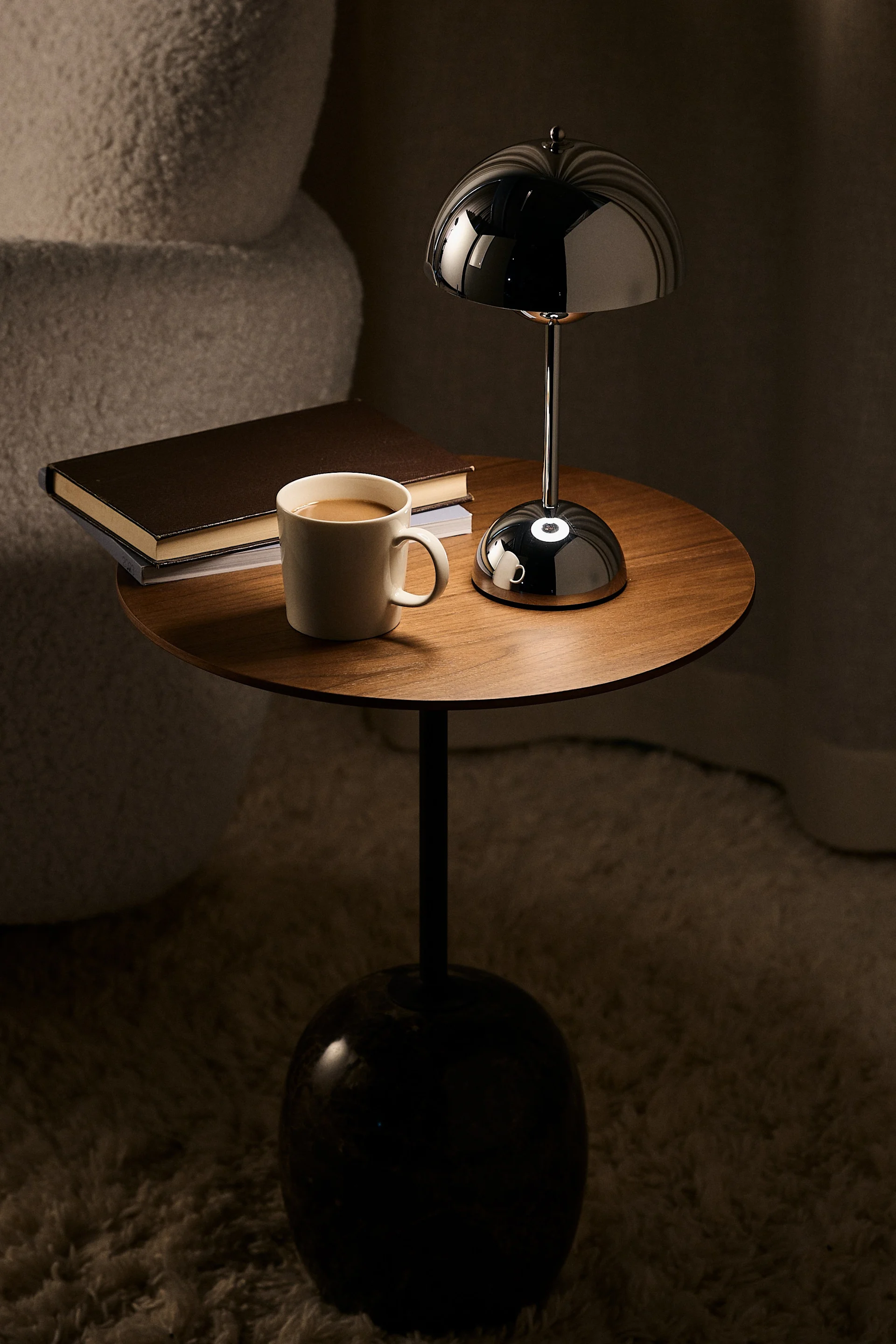 Round wooden table with a glossy black mushroom-shaped lamp, white coffee mug, and books in a dimly lit room.