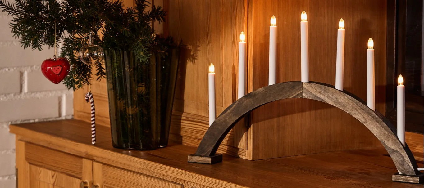 A dark wooden candle arch with seven glowing white electric candles next to a vase of pine branches with a red heart ornament and a candy cane, all on a wooden cabinet.