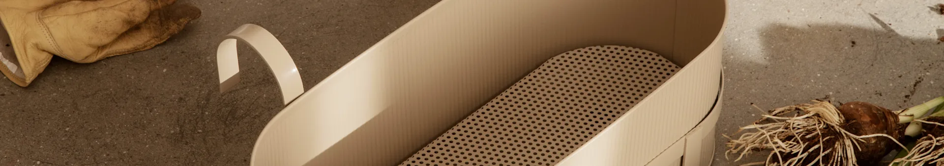 Worn gardening gloves, a light beige planter with a perforated insert, and plant bulbs on a concrete surface.