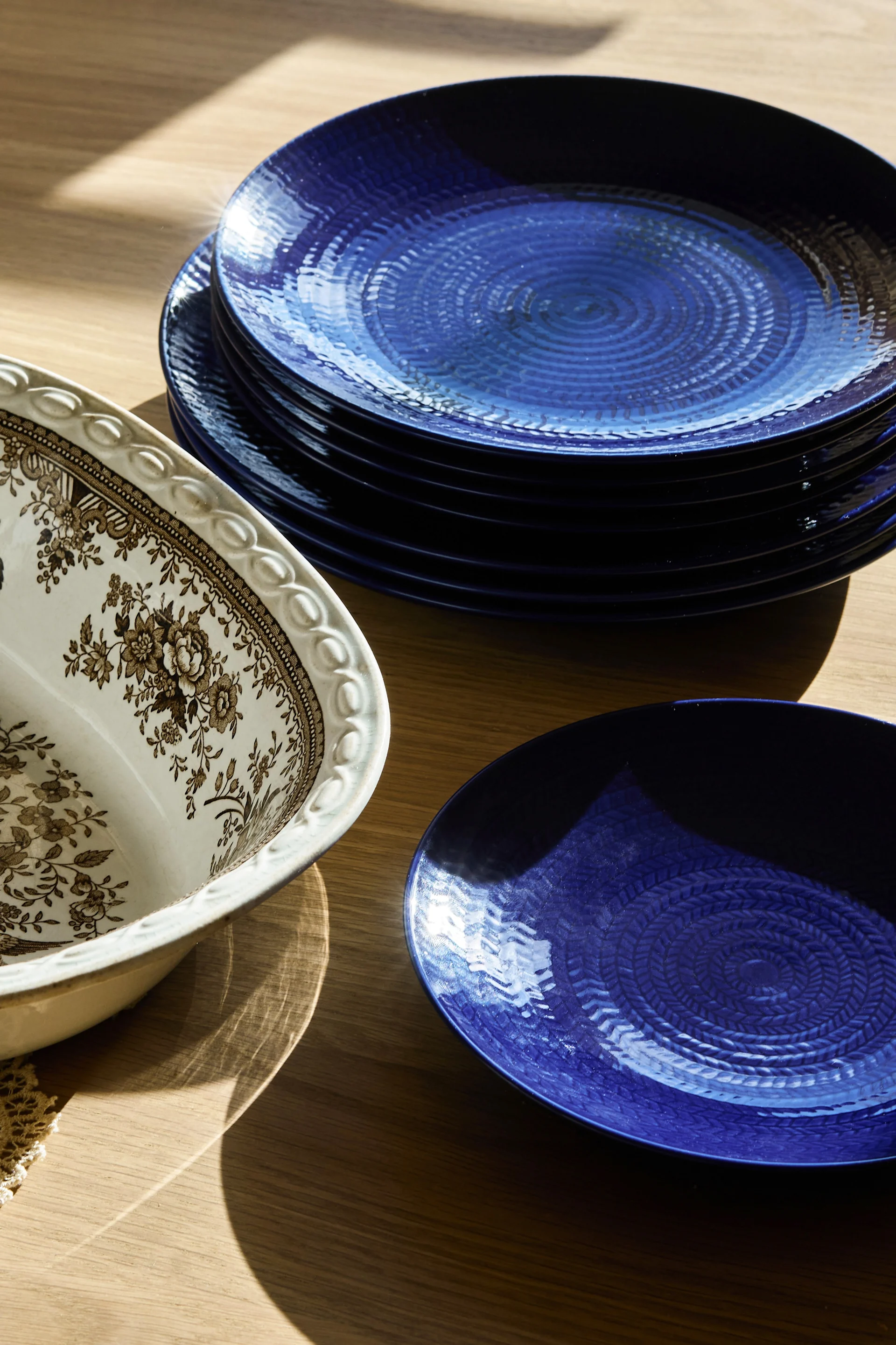 A stack of dark blue, textured plates with a white, brown, and cream floral pattern serving bowl on a wooden table.
