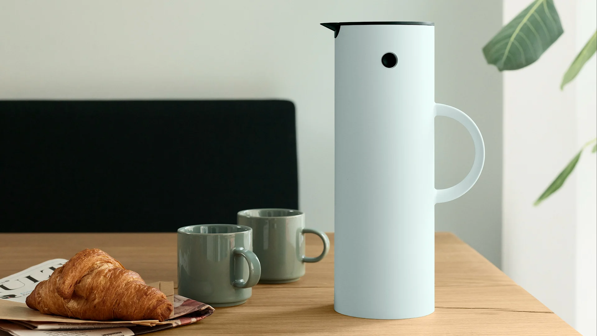 Breakfast setting with a light blue thermal carafe, two grey-green mugs, and a croissant on a wooden table.