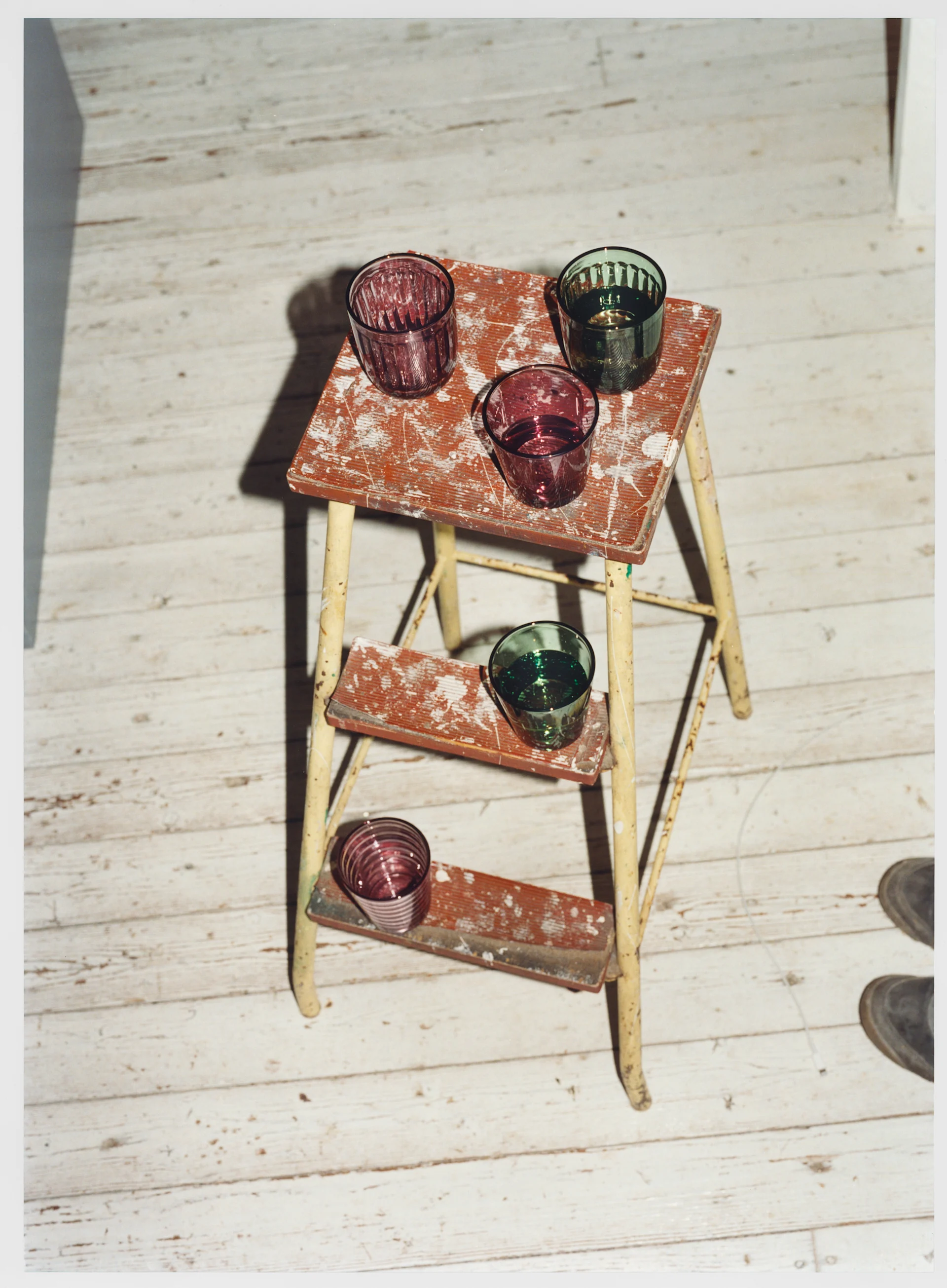 Five ribbed glass tumblers in purple and dark green, arranged on a rustic, worn red three-step ladder on a light wooden floor.