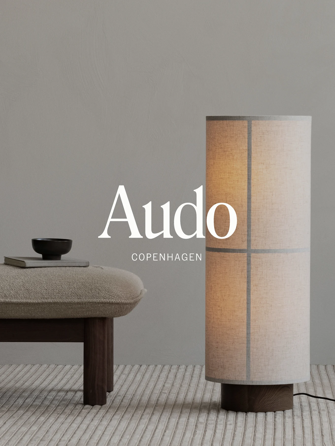 Floor lamp with a cylindrical fabric shade and wood base next to a textured bench with a book and bowl, all on a ribbed rug.