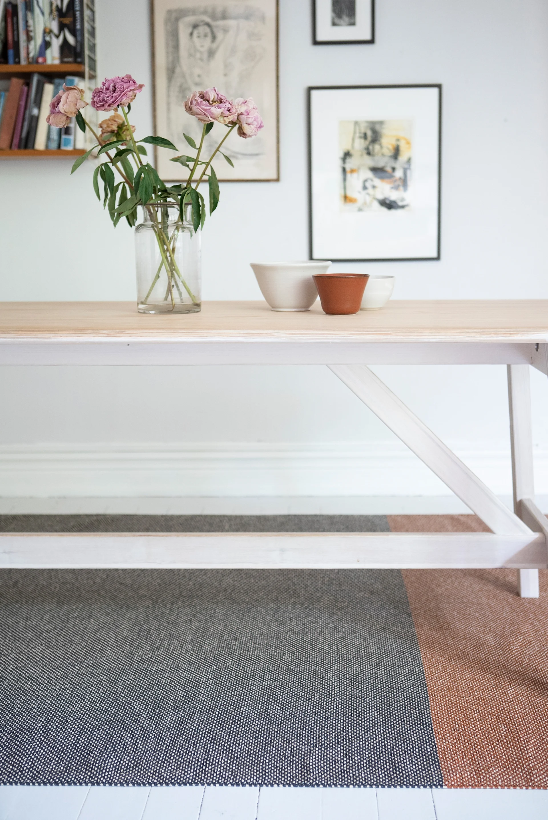 Dining table with fading pink peonies in a vase, ceramic bowls, and a color-block rug.