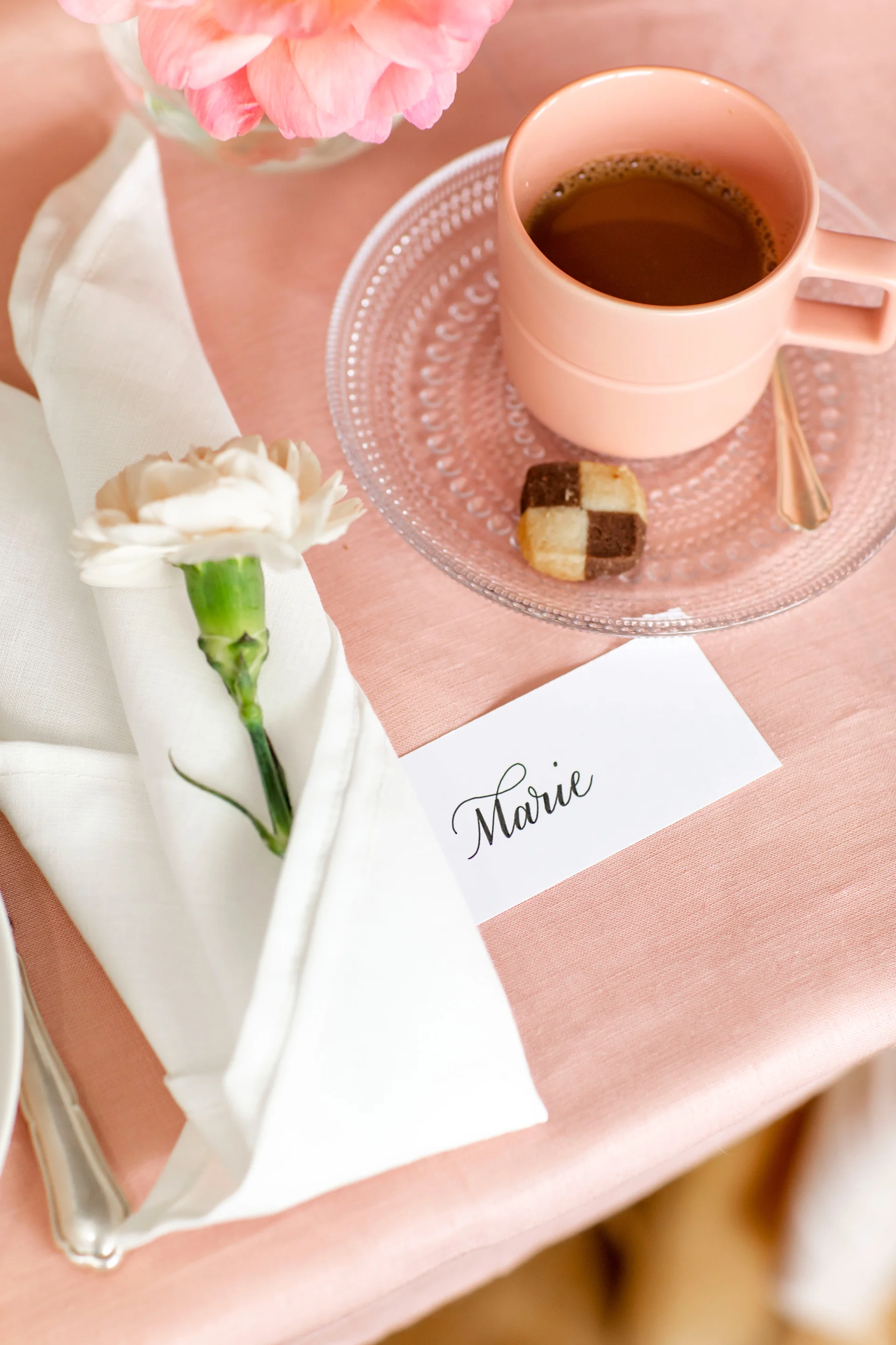 Elegant table setting: pink coffee, two-toned cookie, white carnation in napkin, 'Marie' place card on pink tablecloth.