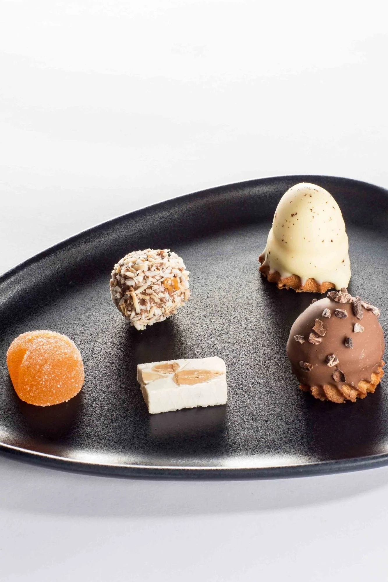 Five assorted gourmet confections on a black plate: white chocolate dome, dark chocolate dome, coconut truffle, orange jelly, and almond nougat.