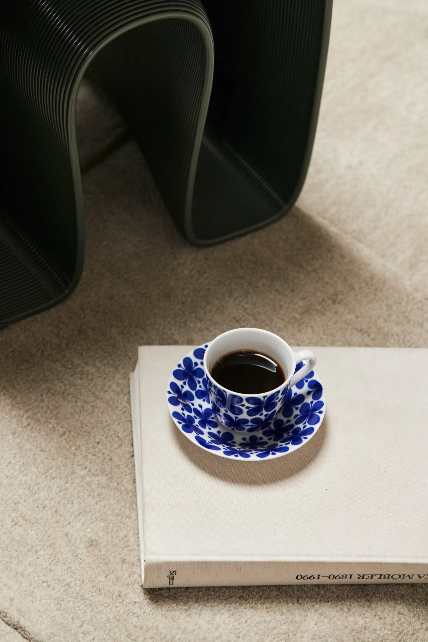 Coffee cup with blue floral saucer on a light book, dark ribbed object in background.