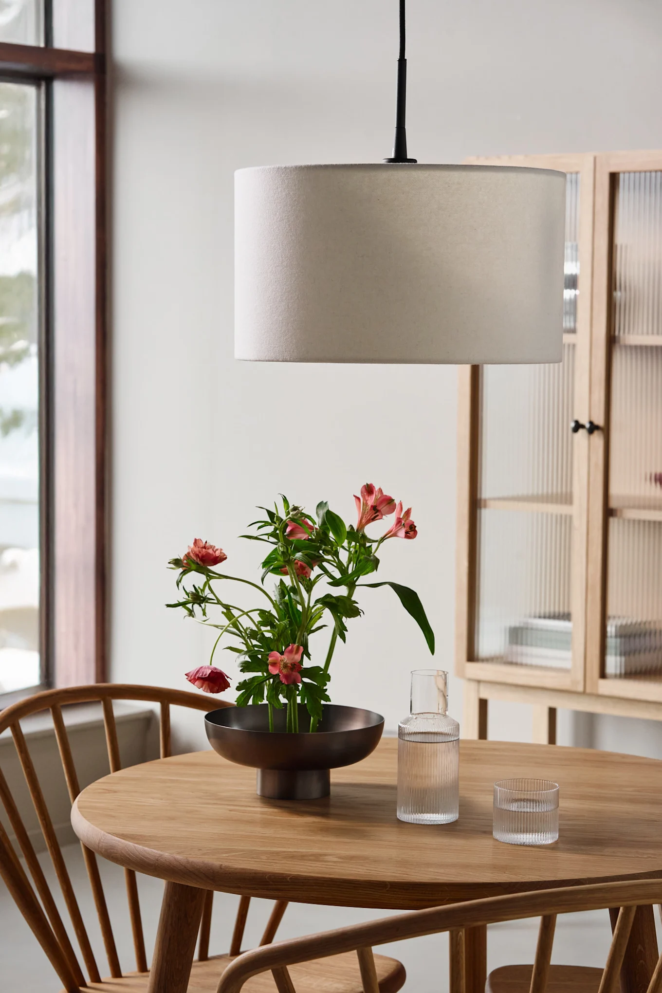 A round wooden table with a bowl of pink flowers, a glass carafe, a tumbler, and a drum pendant light.