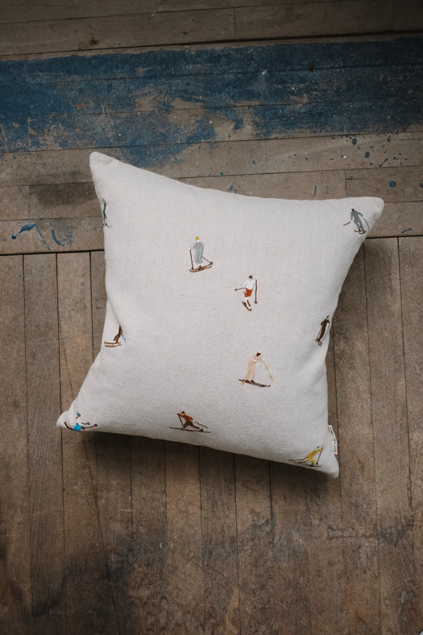 A light-colored throw pillow with embroidered skiers in various poses on a rustic wooden floor.