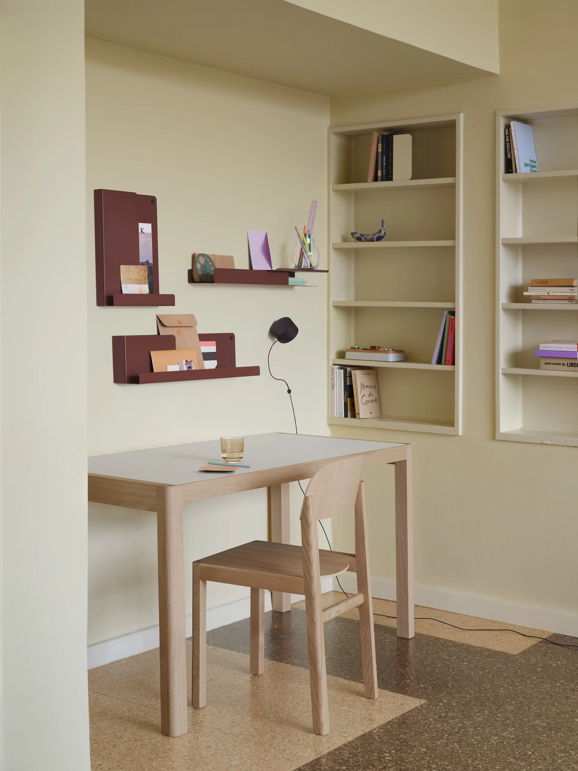 A minimalist home office with a light wood table, chair, and three reddish-brown wall shelves. Built-in bookshelves filled with books are on the right.