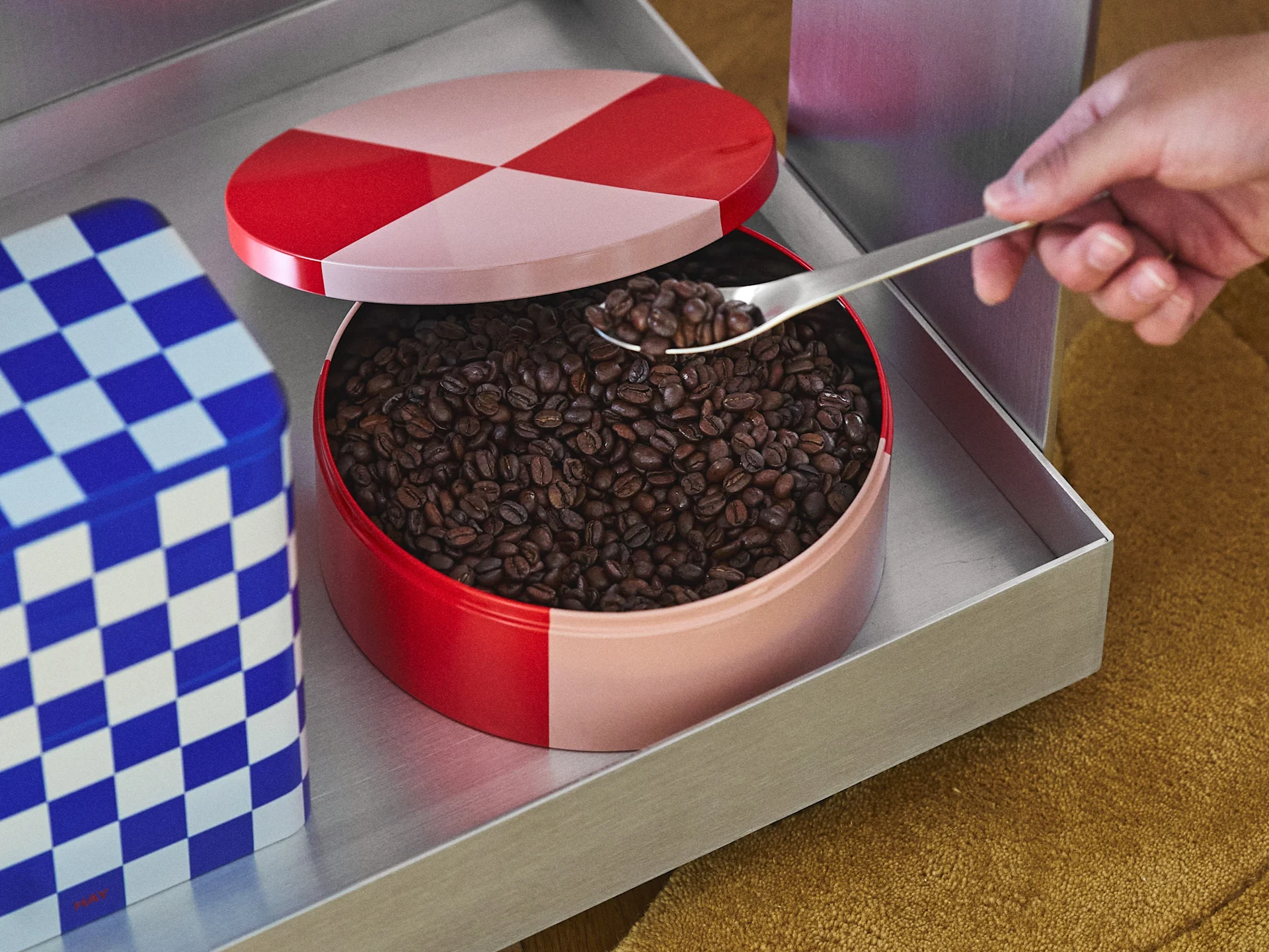 Hand scooping coffee beans from a red and pink checkered tin with a spoon, a blue checkered tin visible to the side.