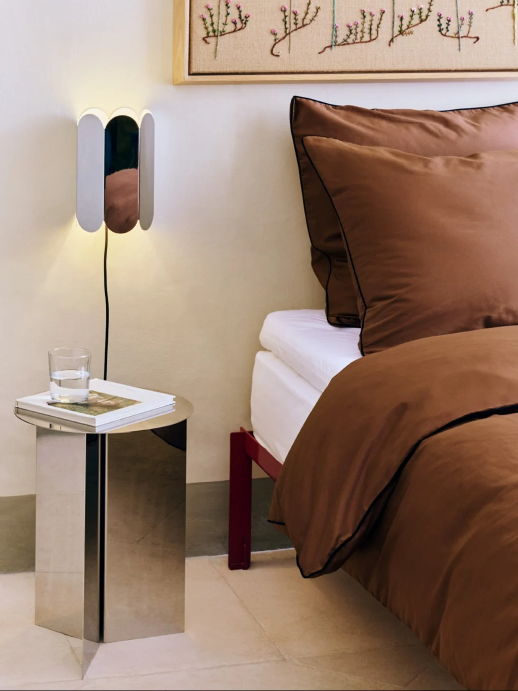 Modern bedroom with a unique wall lamp, metallic side table, and a bed with rich brown bedding.