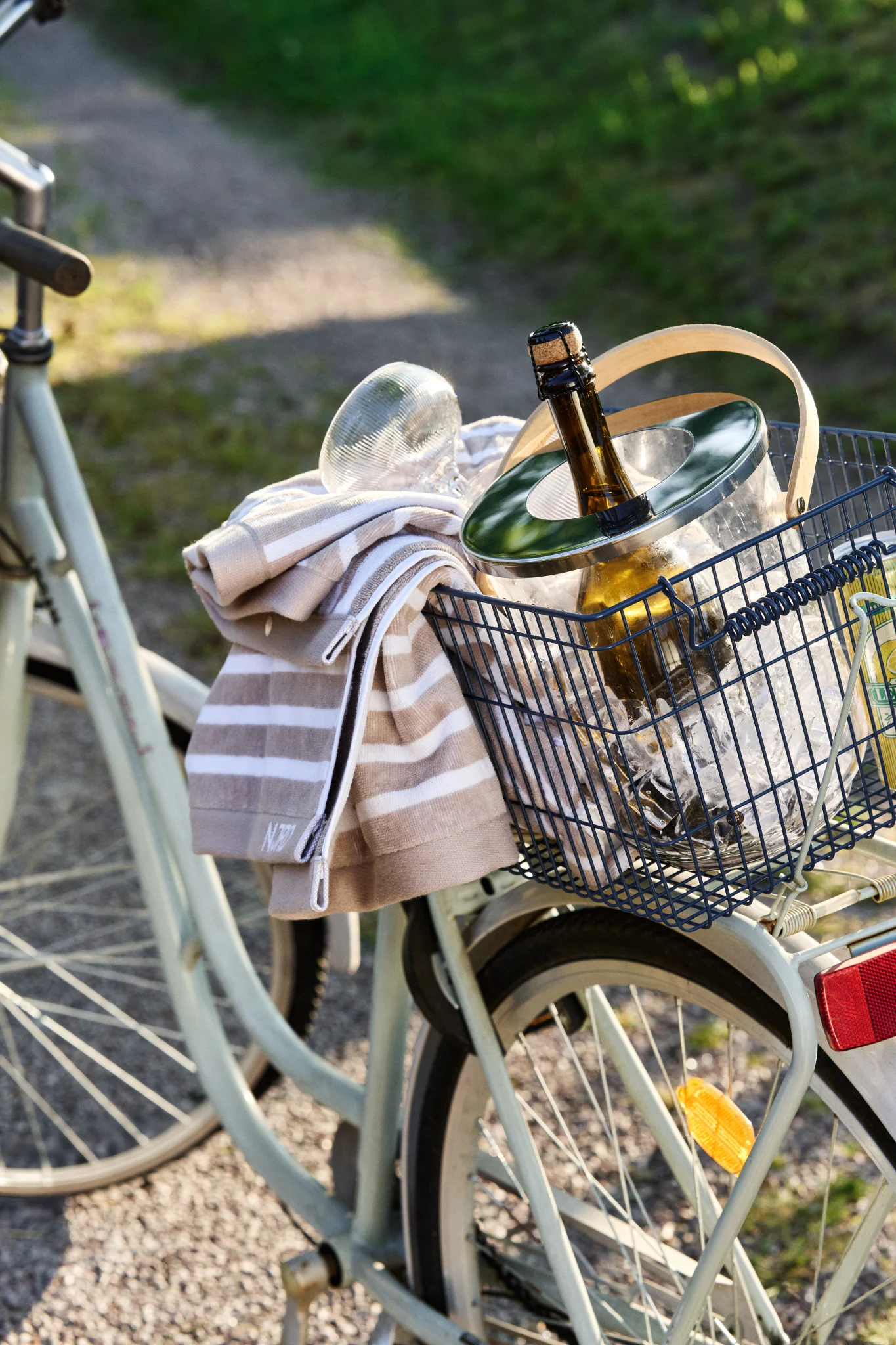 A bicycle basket holds an ice bucket with a chilling bottle and a striped towel on a sunny day.