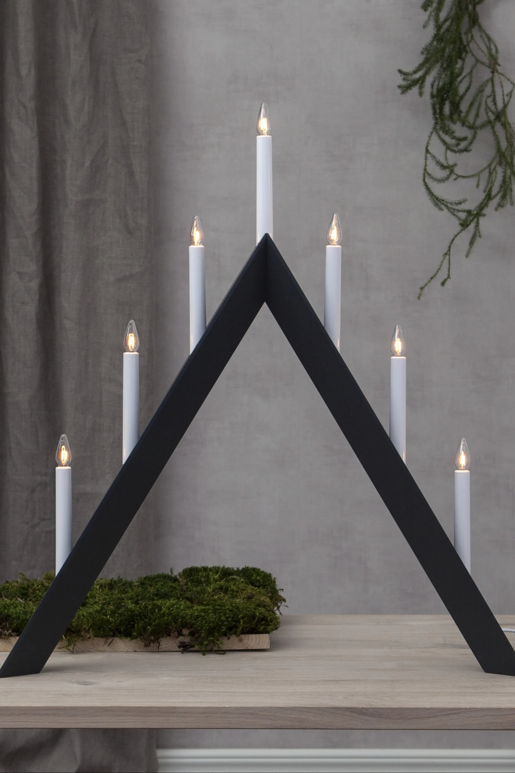 A white, triangular candle holder with seven lit electric candles sits on a white surface, with a blurred Christmas tree in the background.