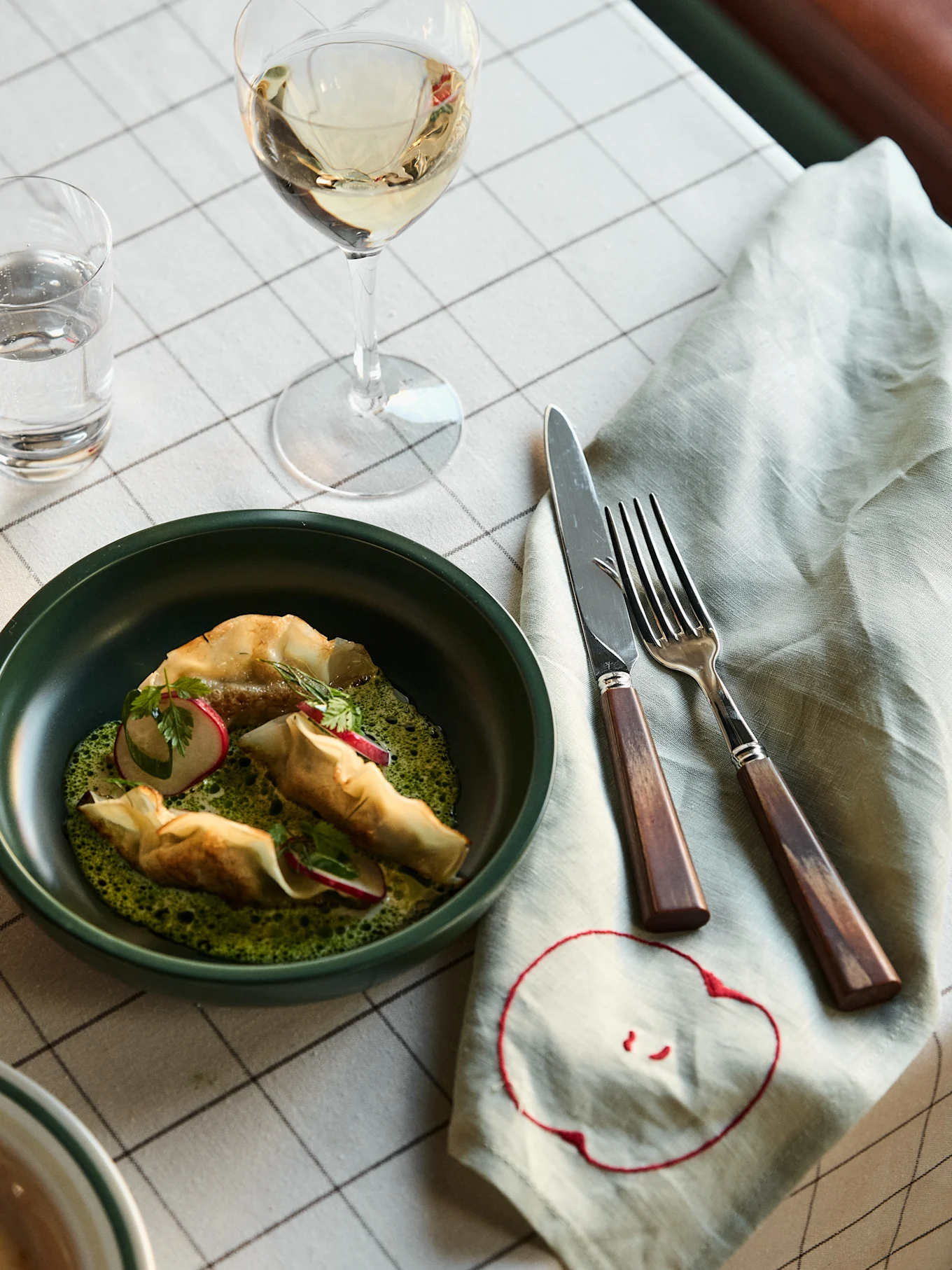 Elegant dish with crispy pastries, green sauce, radish, and herbs in a dark bowl, served with white wine on a checkered tablecloth.