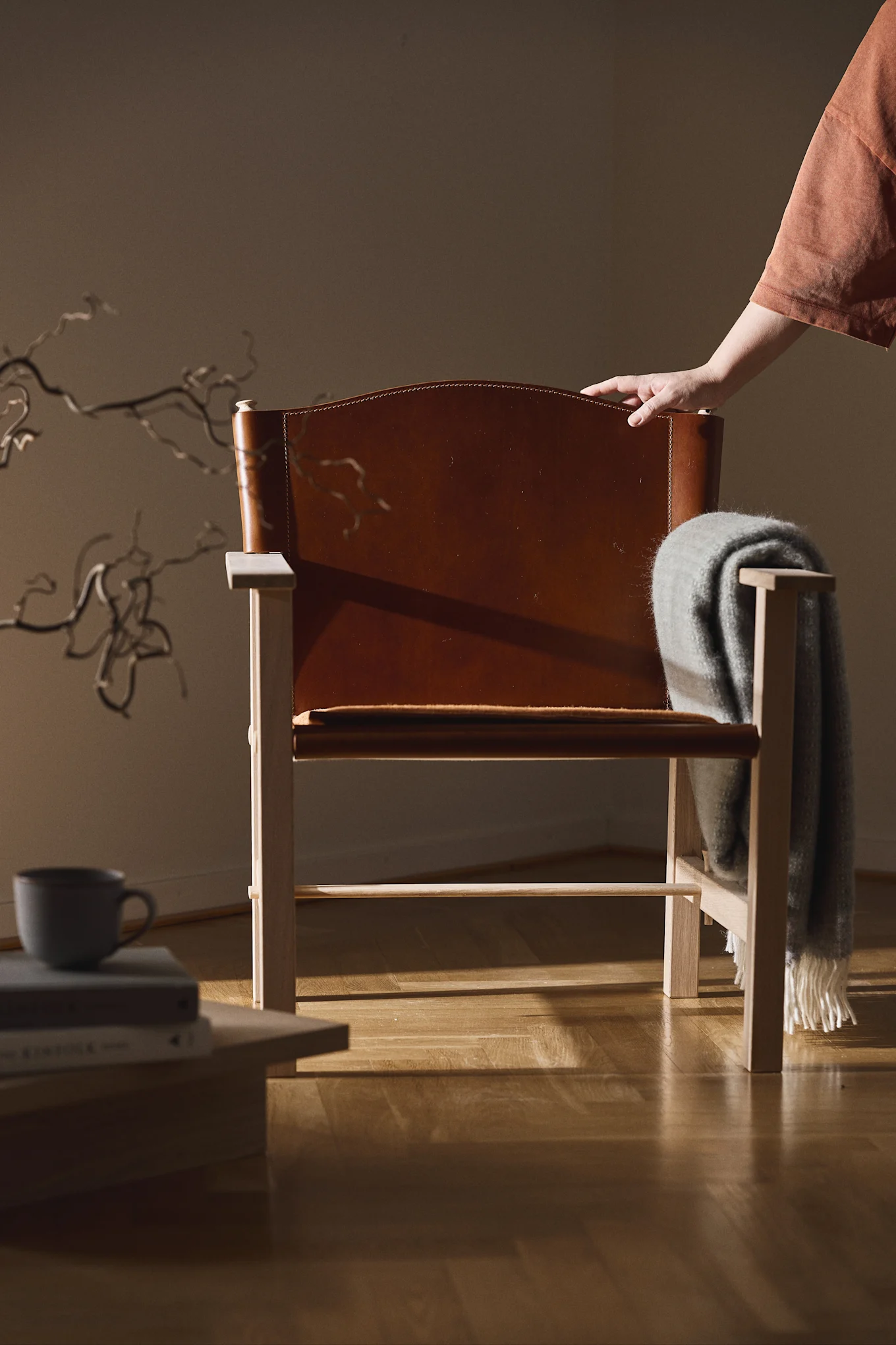 Light wood and brown leather chair, a grey blanket over armrest, and a hand on its back. A mug sits on books.