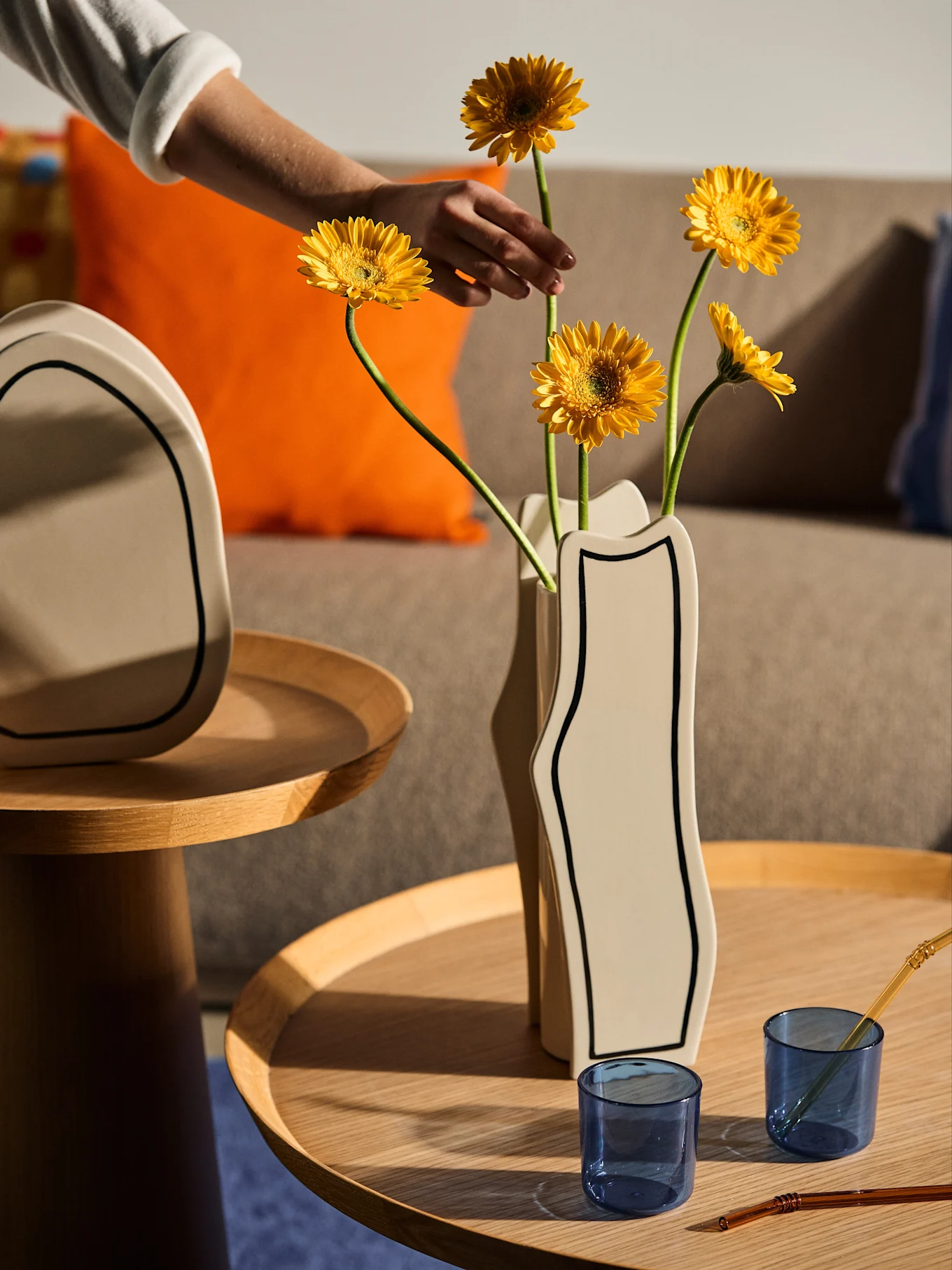 A hand arranges yellow daisies in a wavy light vase on a wooden table with blue glass cups and straws.