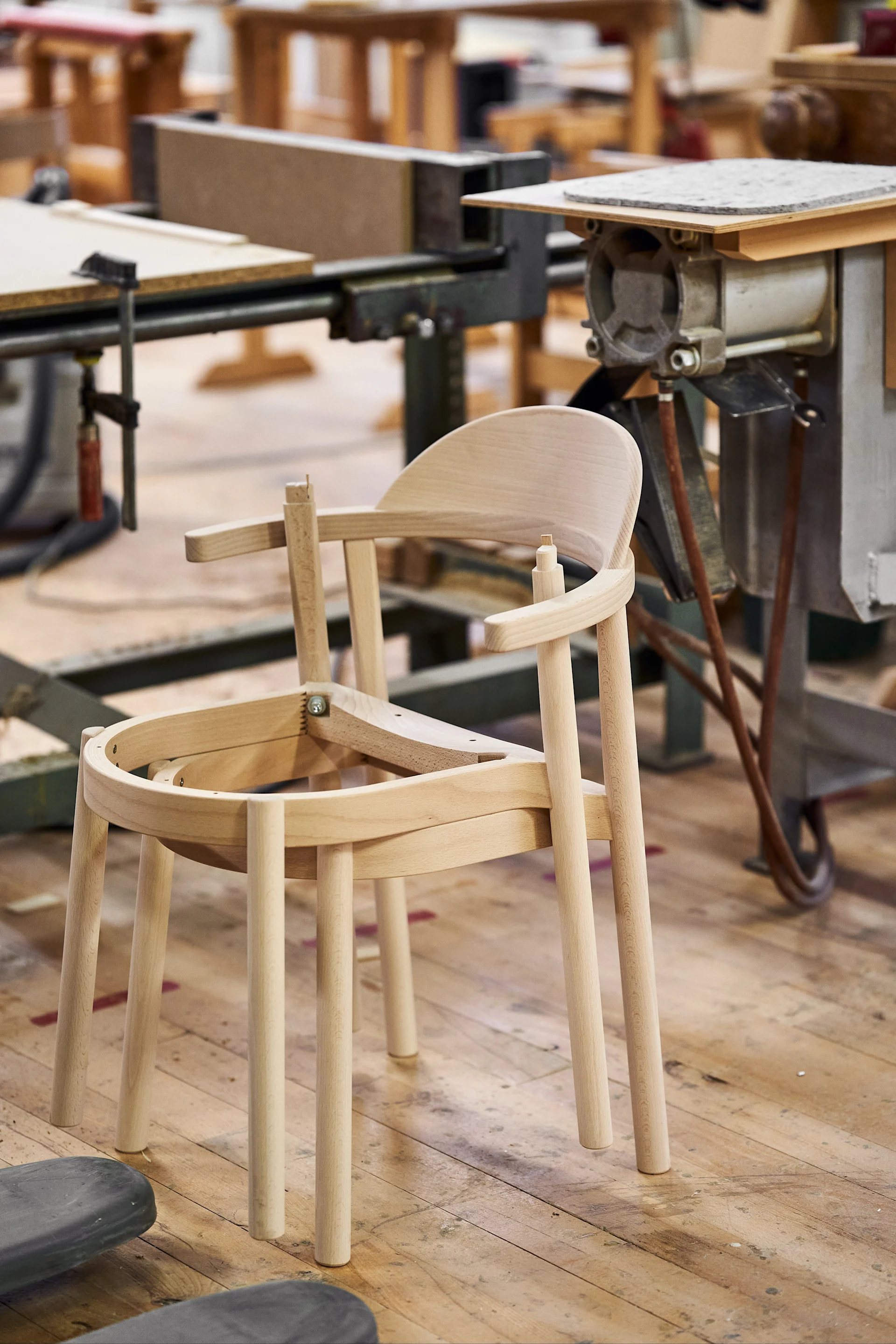 An unfinished light wood chair with a curved back and armrests, standing on a workshop floor with machinery in the background.