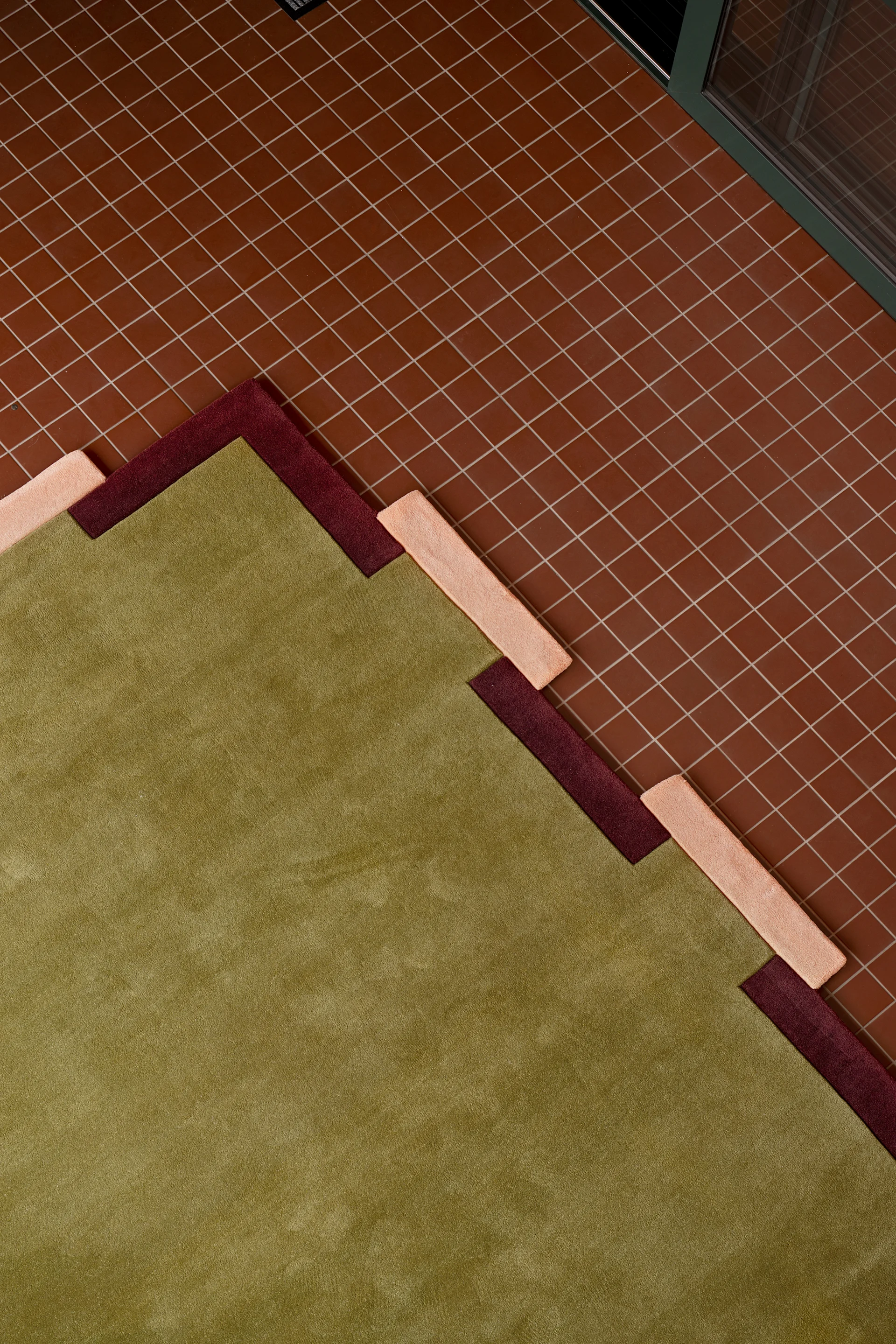 A geometric rug in green, maroon, and peach with a stepped pattern, laid on a reddish-brown tiled floor.