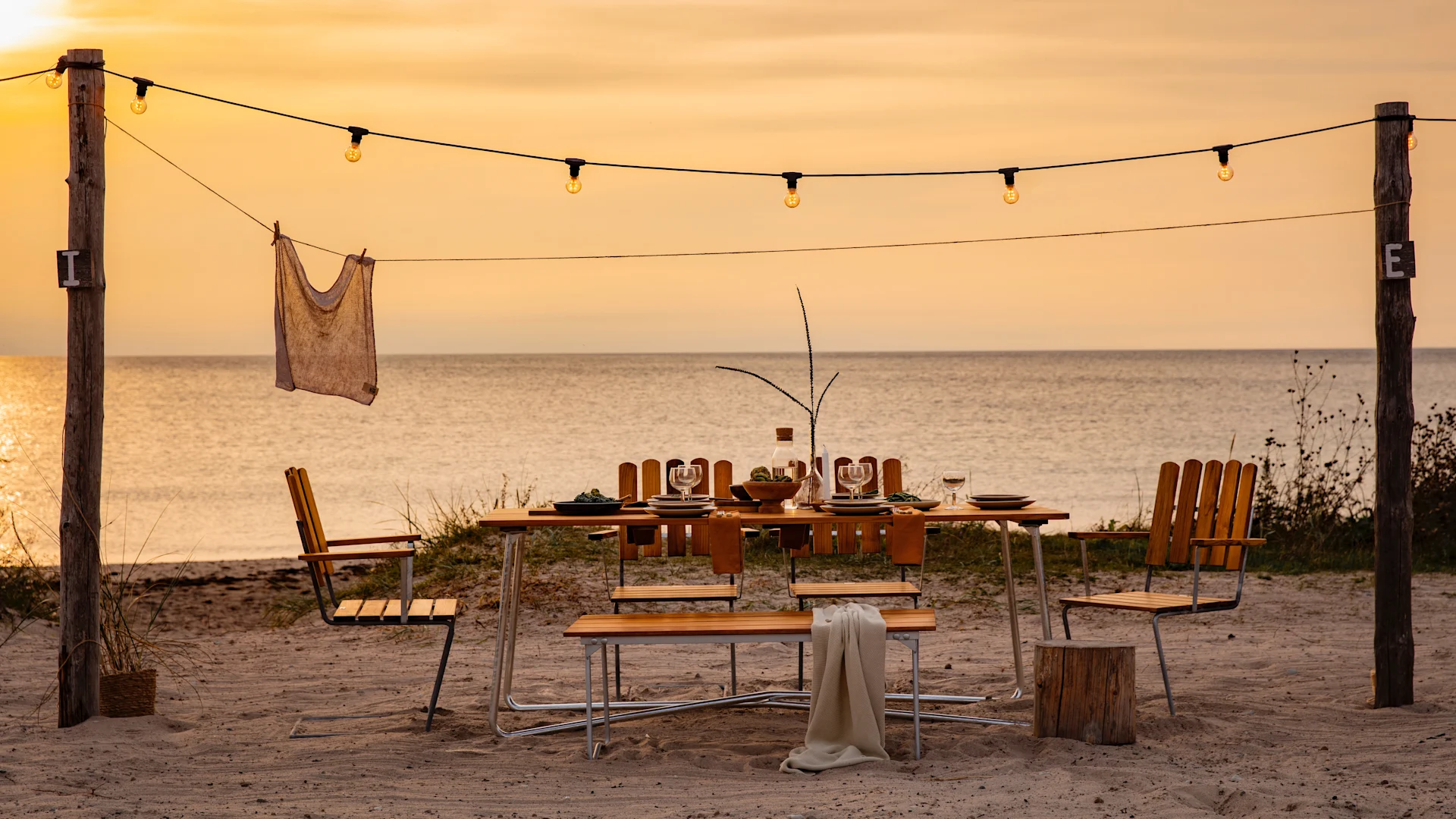 A beautifully set dining table with chairs and string lights on a sandy beach at sunset, with the ocean in the background.