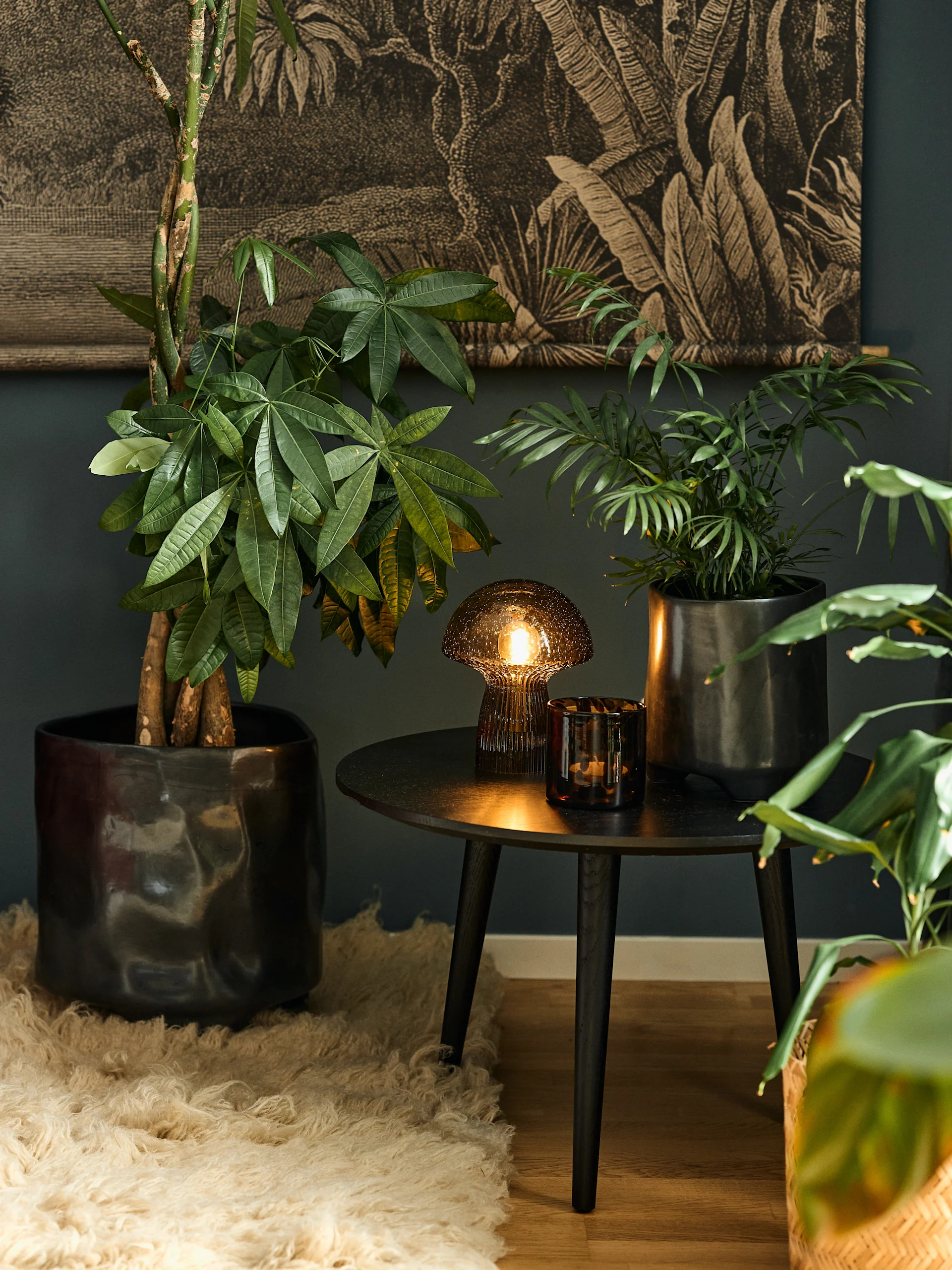 Cozy room with dark walls and tropical plant wallpaper. A large plant, a glowing table lamp, and a candle on a side table.