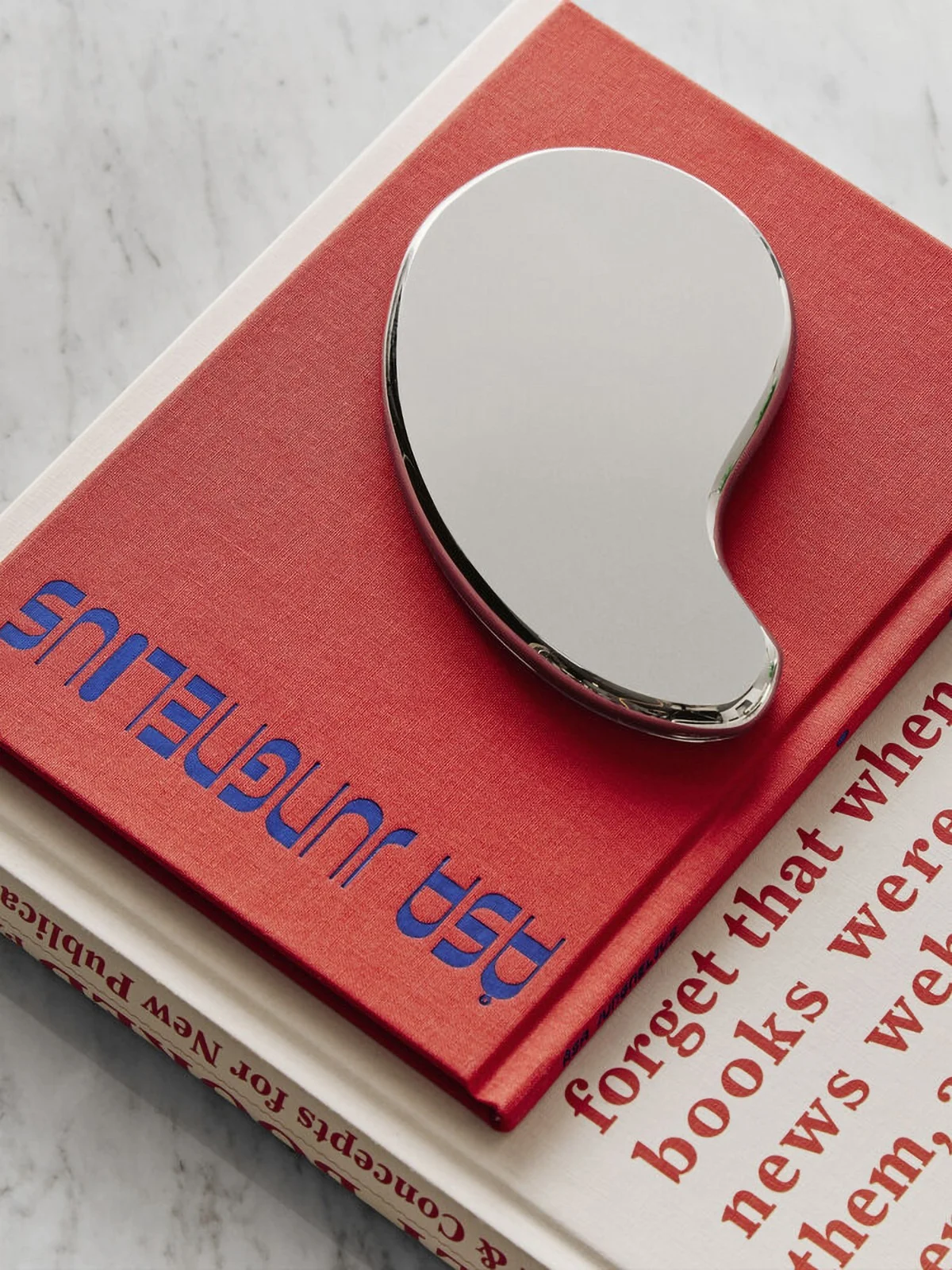 A shiny silver gua sha tool rests on a red book with blue text, atop an off-white book, all on a marble surface.