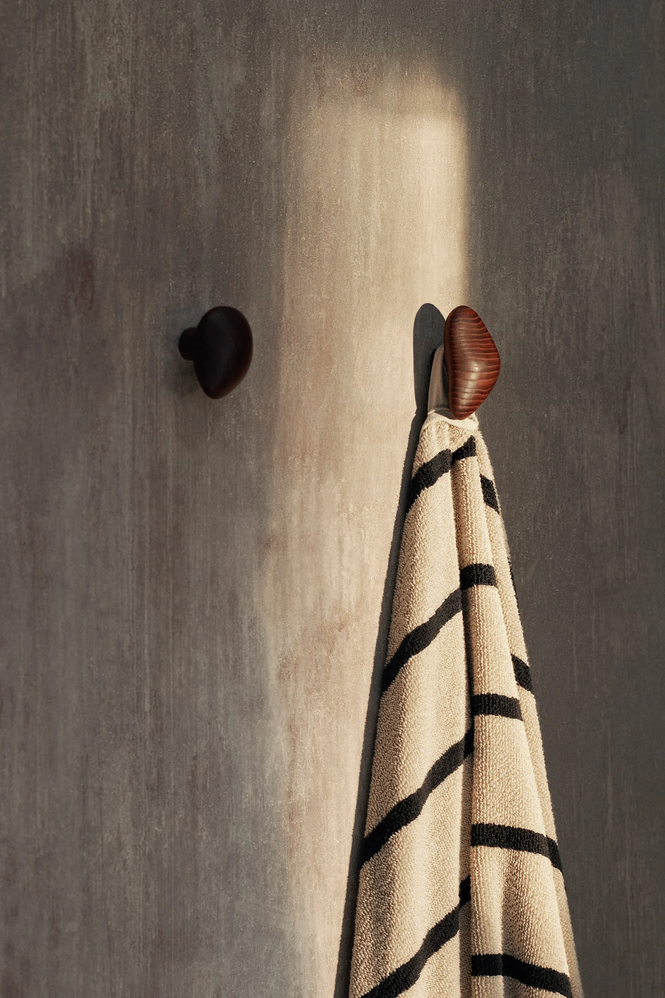 Two wooden wall hooks on a textured wall, one holding a beige towel with black stripes, highlighted by sunlight.