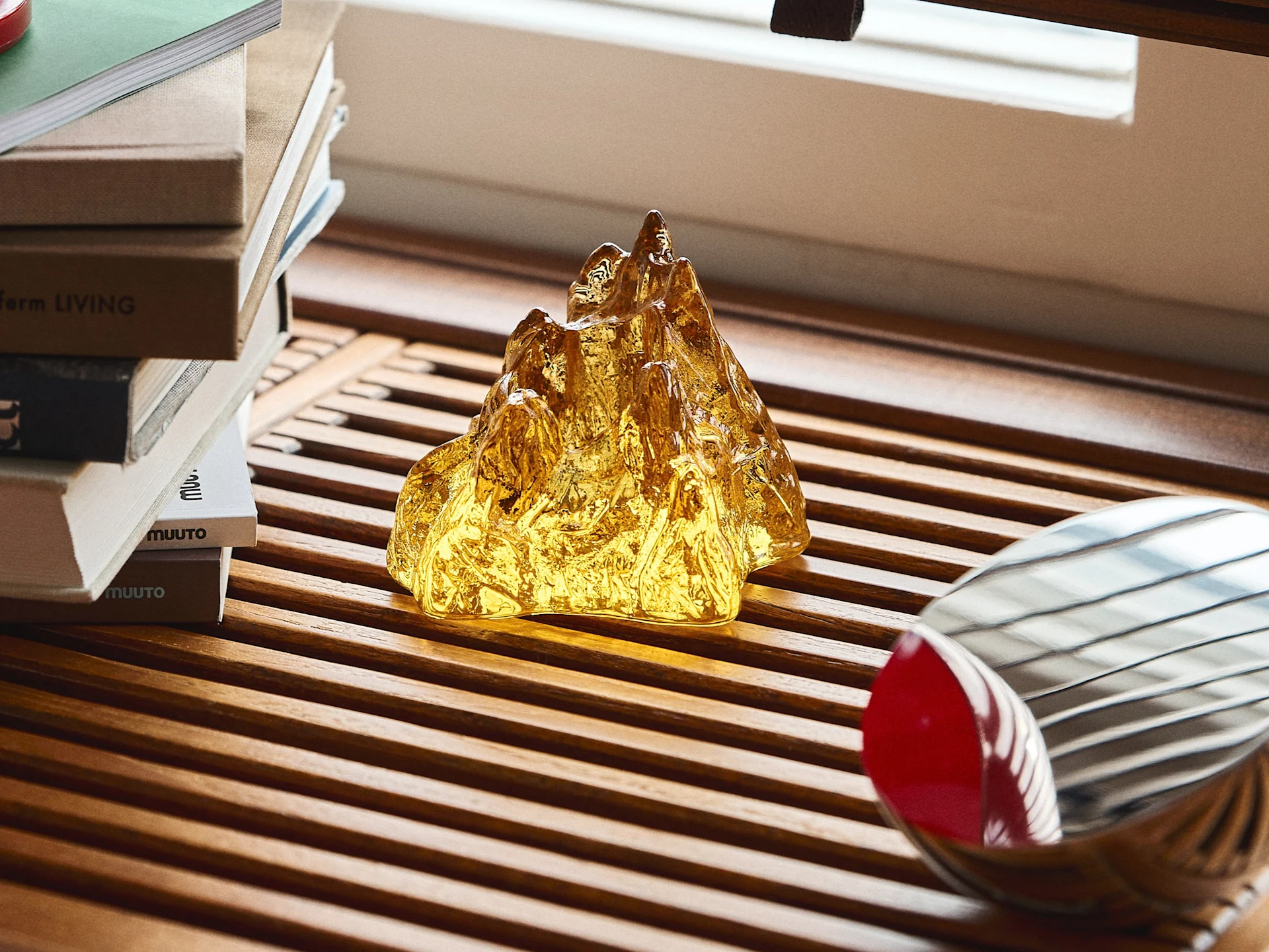 Golden mountain-like sculpture on a wooden slatted table next to a stack of books and a metallic object.