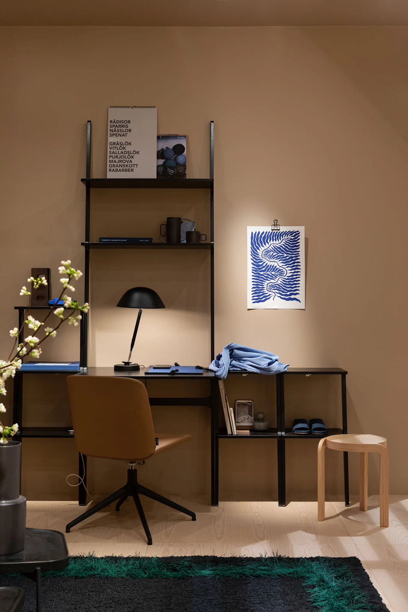 A modern home office with a black shelving unit, a brown swivel chair, a black desk lamp, blue art, and a green-striped rug.